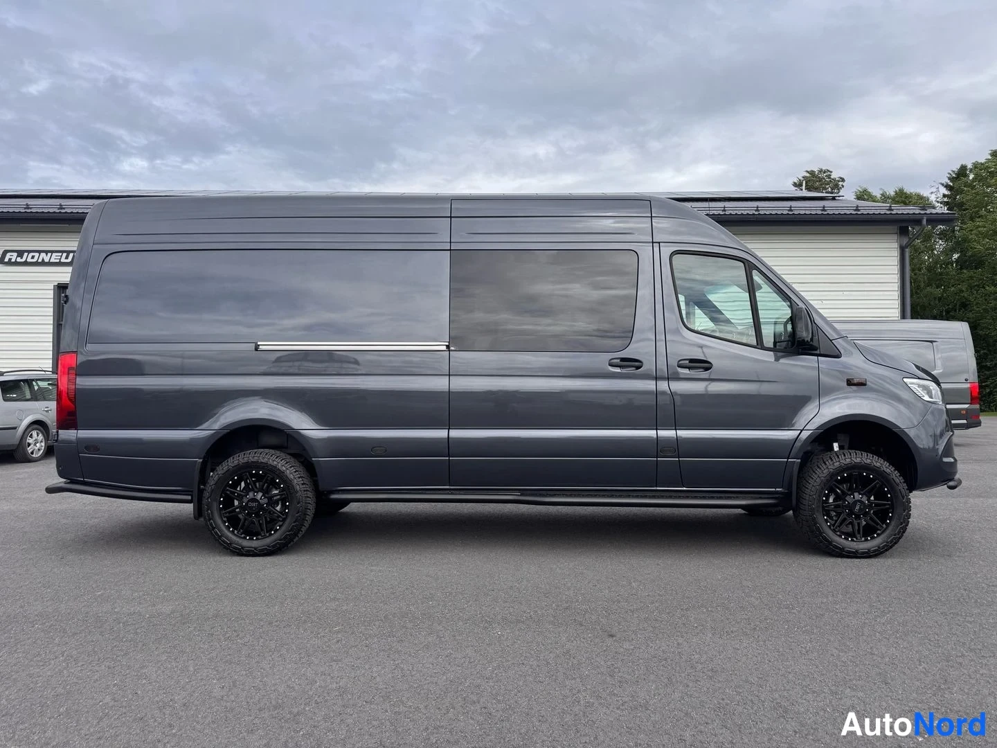 Sivulta: Sivukuva tummanharmaasta Mercedes-Benz Sprinter -pakettiautosta pitkillä mustilla vanteilla.