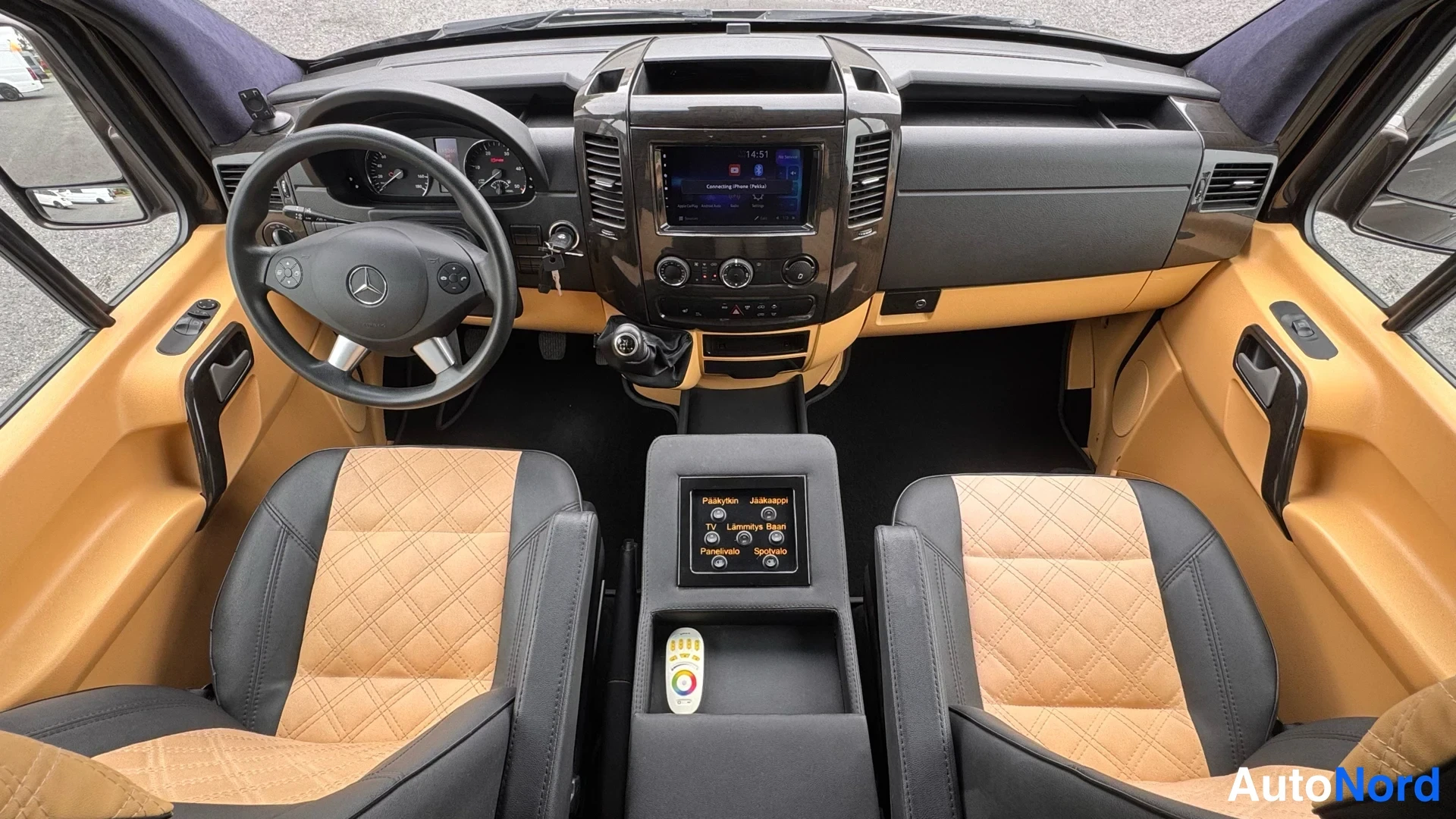 Edestä: Ajo-osaston sisäankaappi: Mercedes-ohjauspyörä, tumma kojelauta, kosketusnäyttö ja kaksi dväkuvioitua musta-beige istuinta.