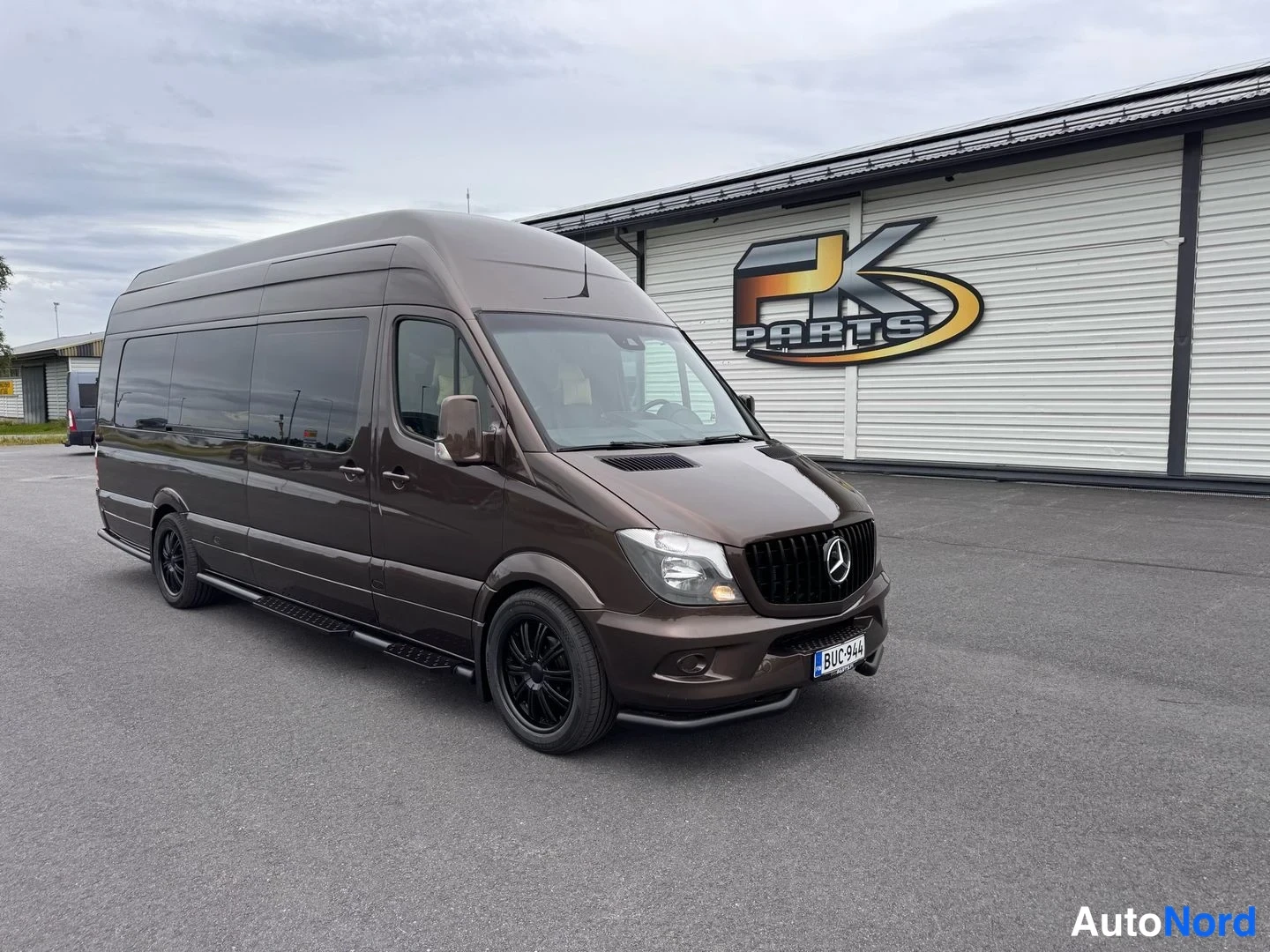Etuviisto, nokka oikealle: Mercedes-Benz Sprinter -pakettiauton etuosasta viistosti otettu kuva, keula oikealla, tummanruskea koripinta, mustat vanteet, taustalla PK Parts -rakennu