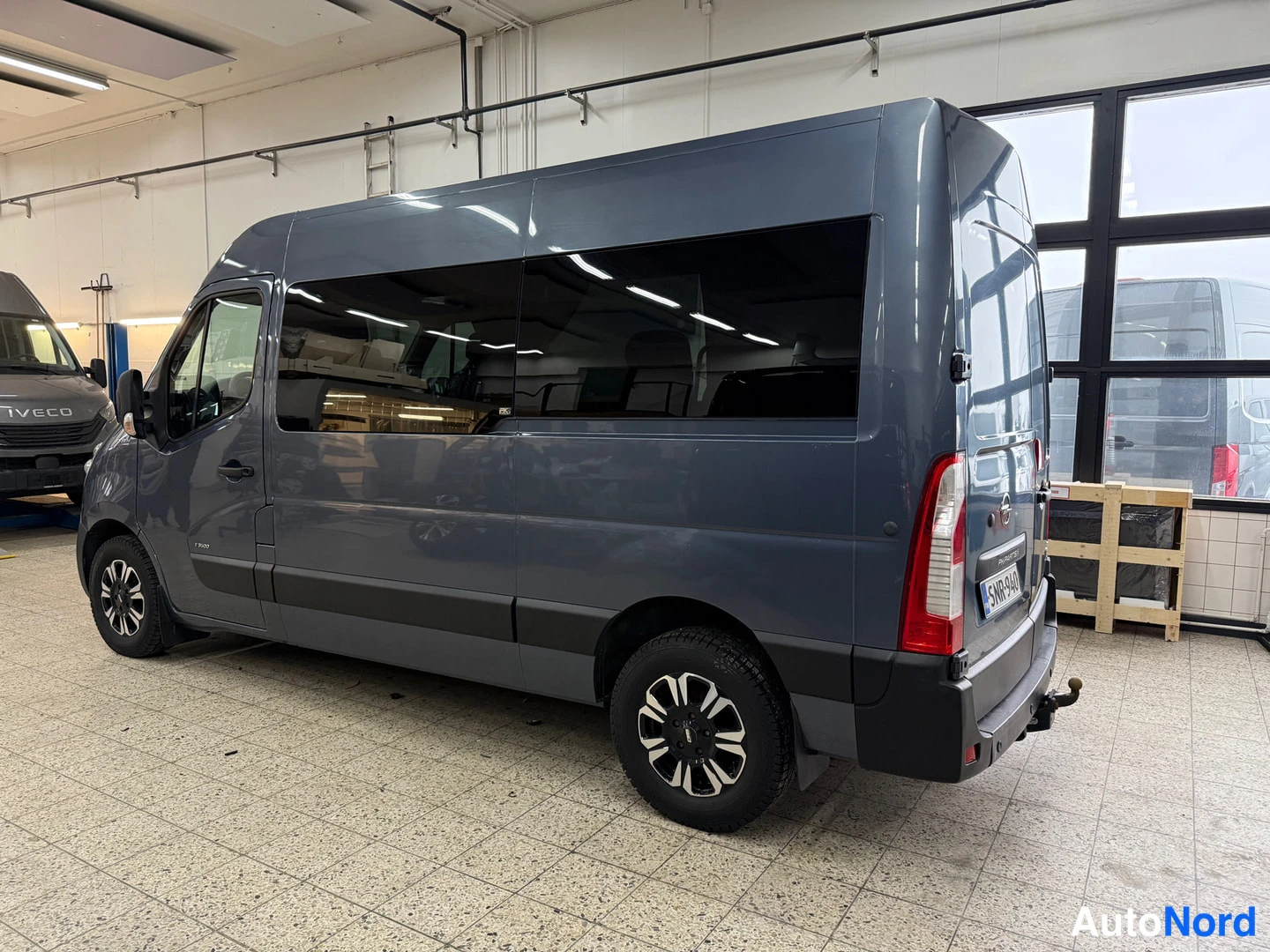 Takaviisto: Tummanharmaan Opel Movano -pakettiauton takaosa ja oikea sivu näkyvät takaosakulmasta sisätiloissa, perävaunukoukku ja rekisterikilpi näkyvissä.