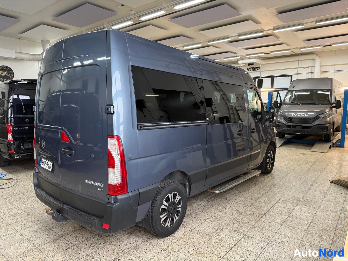 Takaviisto: Tummanharmaan Opel Movano -pakettiauton takaosa ja sivu takaosakulmasta sisätiloissa, sivuille asennettu astinlaudat ja perävaunukoukku näkyvissä.