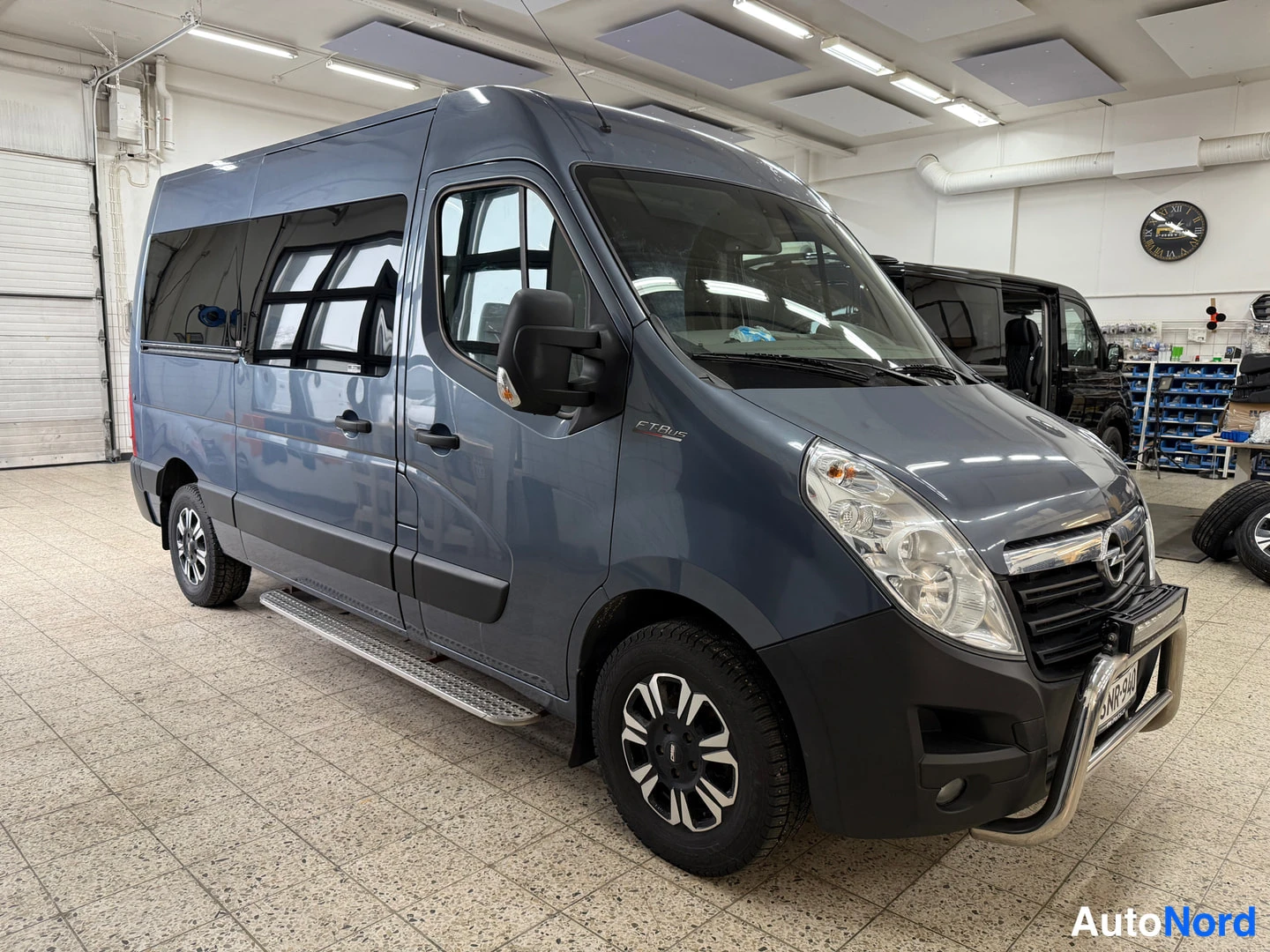 Etuviisto, nokka oikealle: Opel Movano -pakettiauto etuviistöstä oikealta, tummanharmaa kori, sisätiloissa kiiltävät lattiat.