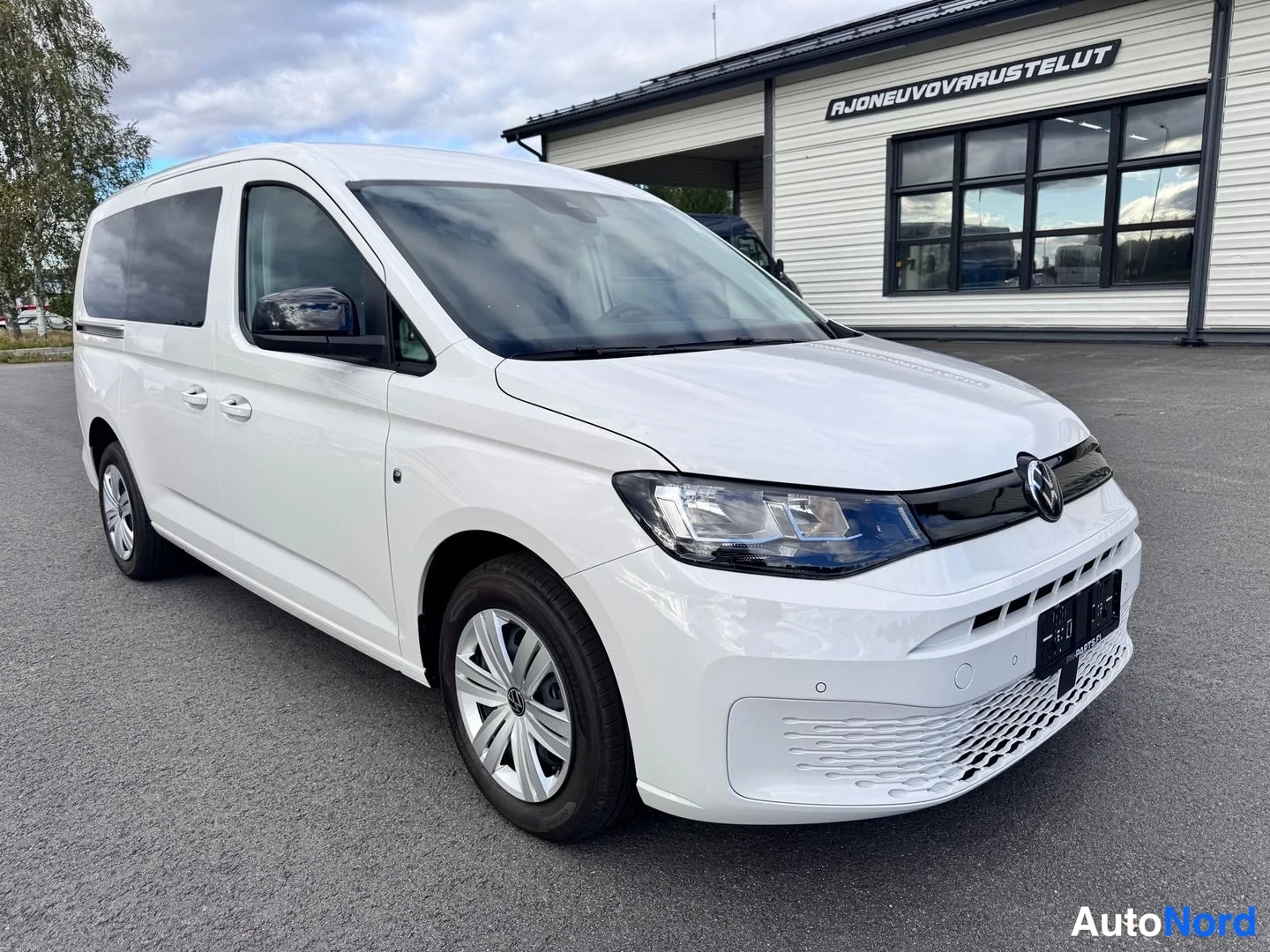 Etuviisto, nokka oikealle: Valkoinen Volkswagen Caddy -pakettiauto etuosaltaan oikeasta etukulmasta kuvattuna paikoitusalueella.