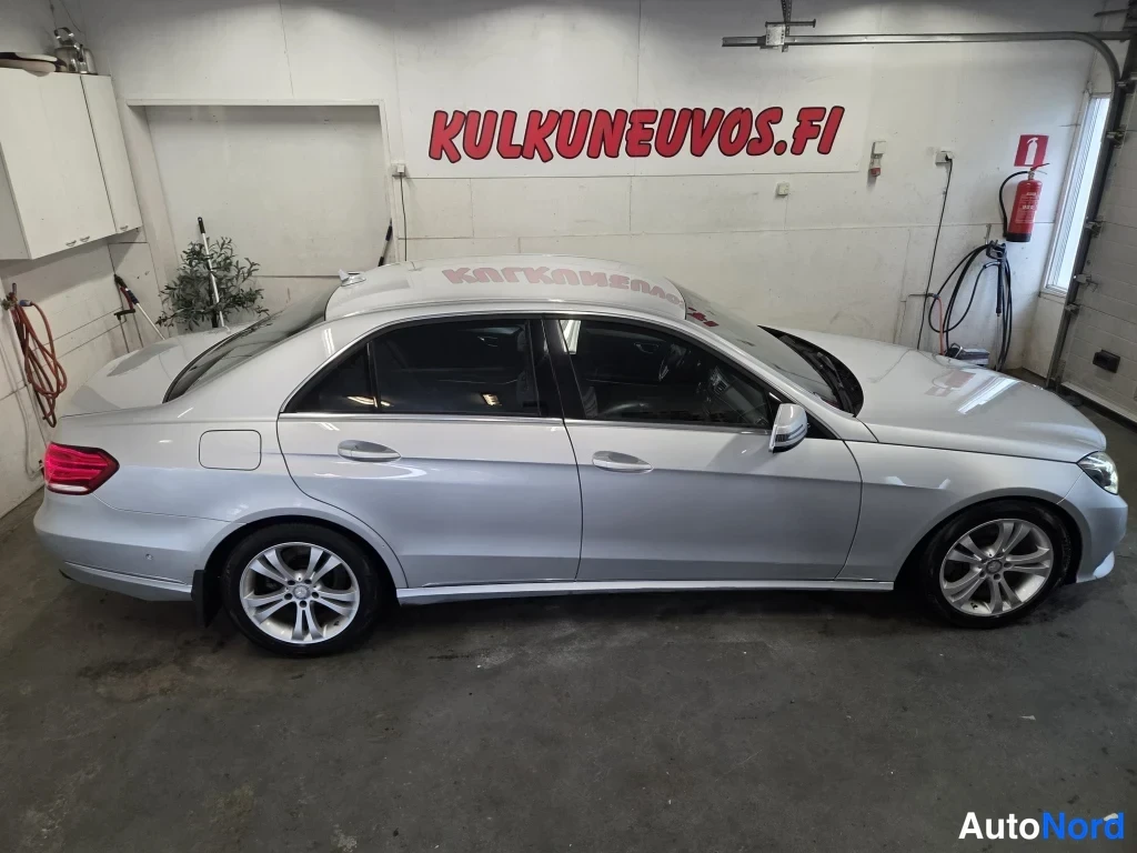 Etuviisto, nokka oikealle: Hopeanvärinen Mercedes-Benz E-sarjan sedan sivulta oikeaan viistosti kuvattuna autonhallissa, seinällä KULKUNEUVOS.FI -teksti.
