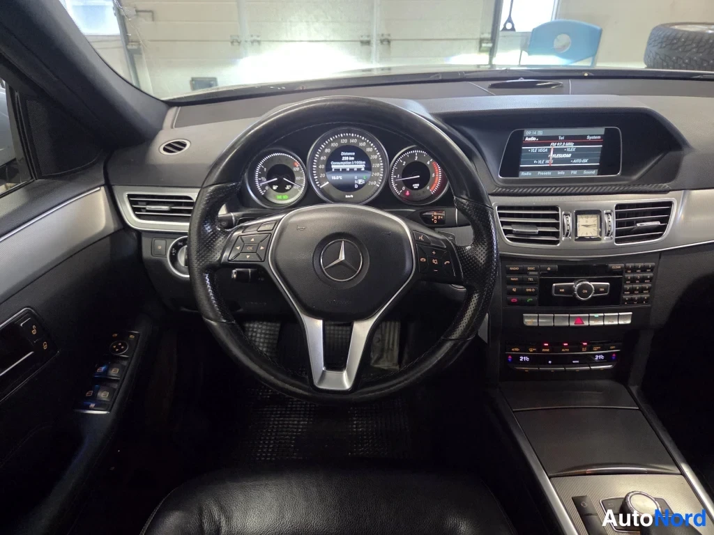 Yksityiskohta: Mercedes-tunnuksellinen ohjauspyörä, mittaristo, keskikonsoli ja infotainment-näyttö, musta sisusta.