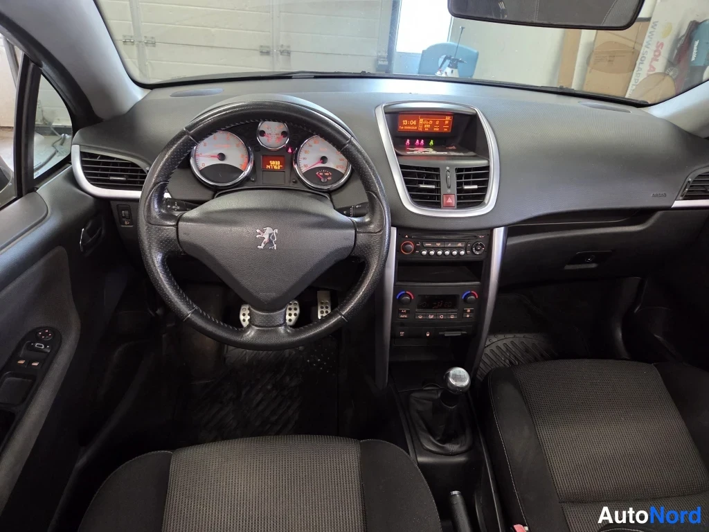 Muu: Peugeot 207:n ohjaamo edestä: ohjauspyörä mittareineen, keskikonsoli ja manuaalivaihteen keilapomo, tumma sisusta.