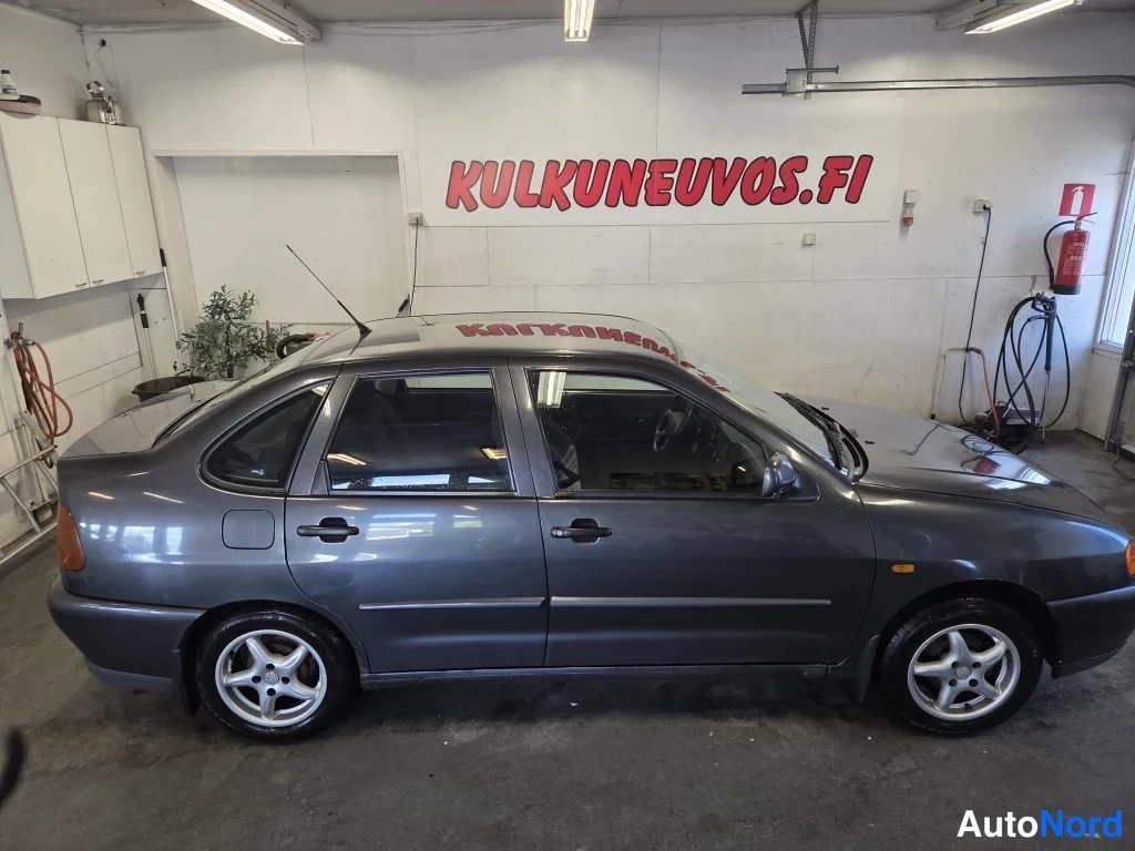 Etuviisto, nokka oikealle: Tummanharmaan Volkswagen Polo Classic -sedanin sivukulmaautokuva sisätiloissa, vasen sivu ja keulan kärki näkyvissä.