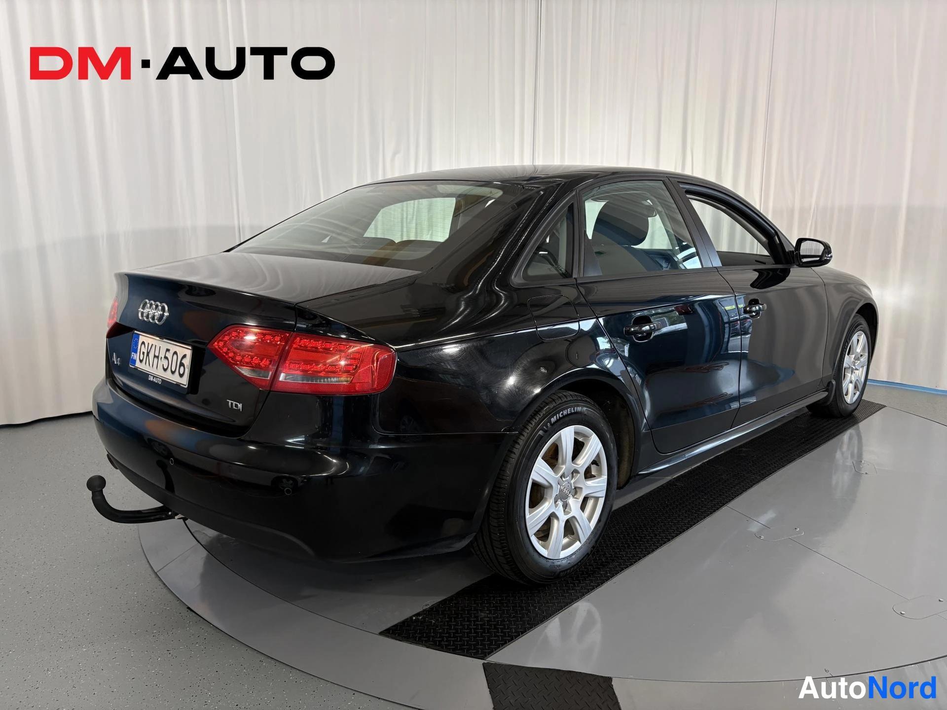 Takaviisto: Audi A4 -sedanin takaosa ja vasen sivu kolmannesnäkymässä; musta koripinta, takavalo, rekisterikilpi GK H-506, A4- ja TDI-merkinnät, perävaunukoukku.