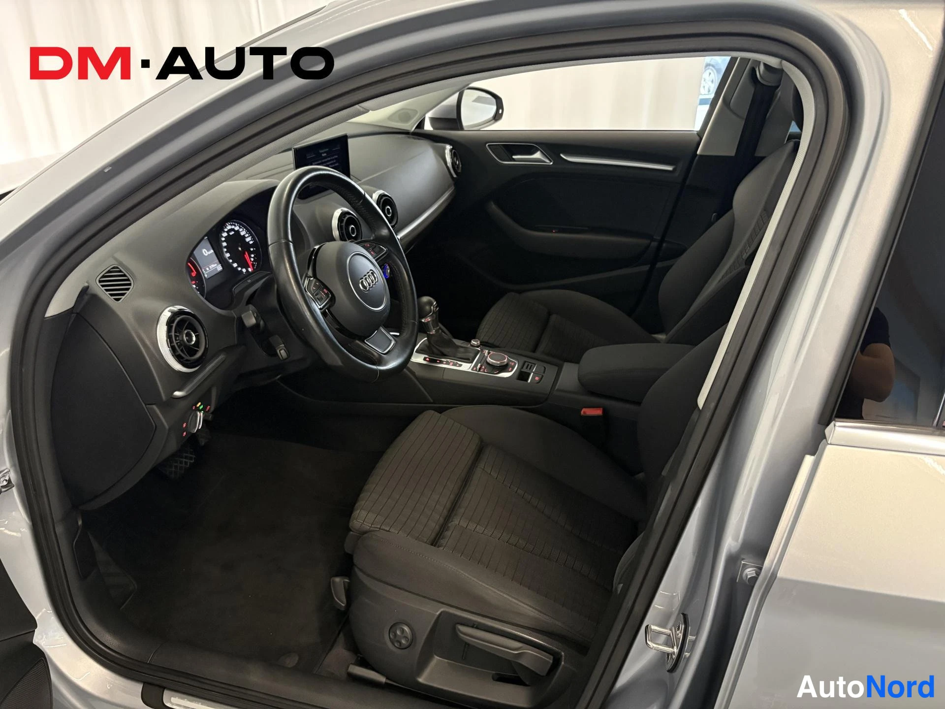 Muu: Audi A3:n ohjaamo avoimesta kuljettajan ovesta kuvattuna: mustat kangasistuimet, multiline-ohjauspyörä, MMI-näyttö ja keskikonsoli.