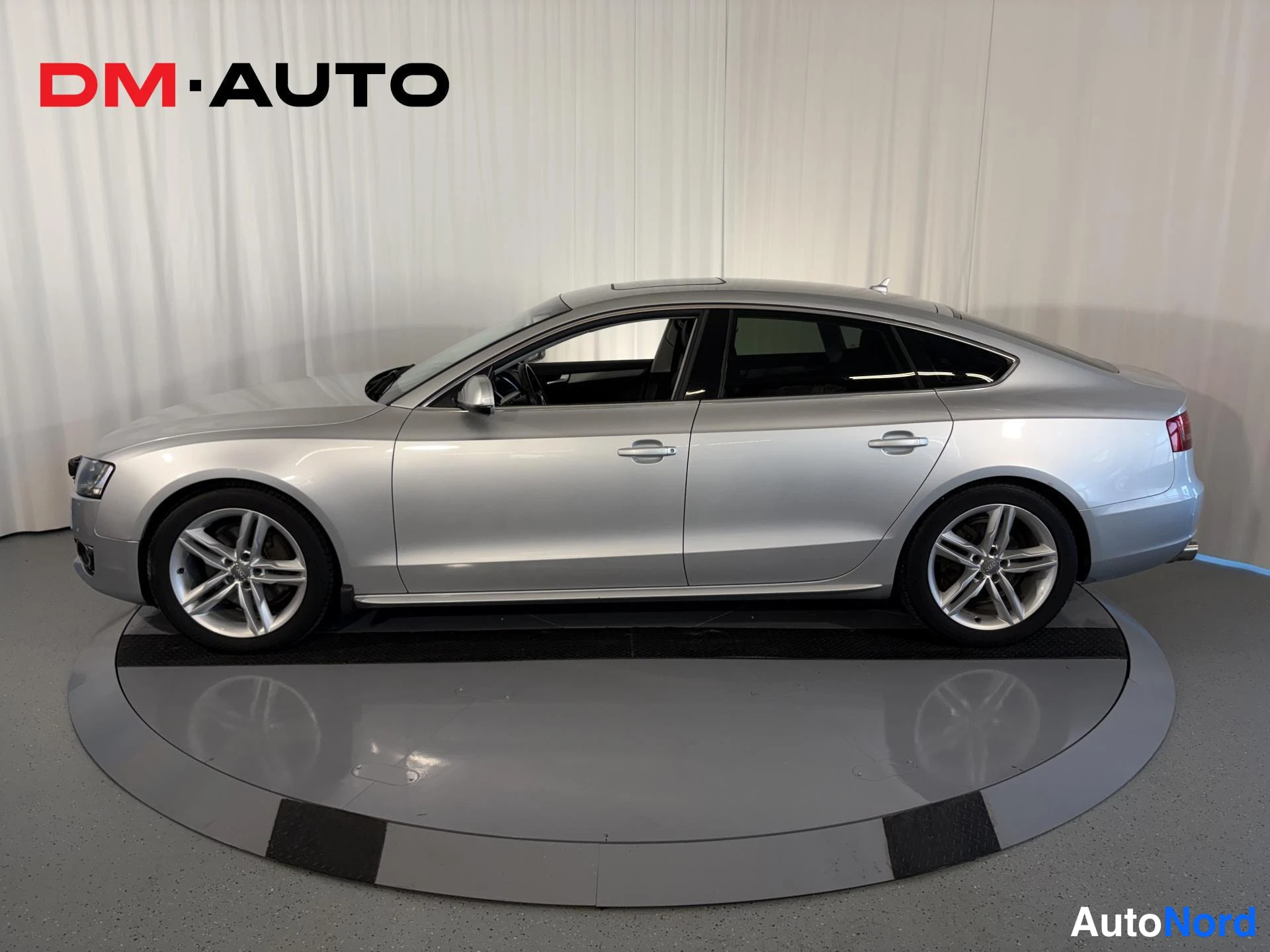 Sivulta: Hopeinen Audi A5 Sportback täsmälleen sivulta vasemmalta kuvattuna studio­pyörätelineessä, viisiovinen runko ja kevytmetallivanteet näkyvät.