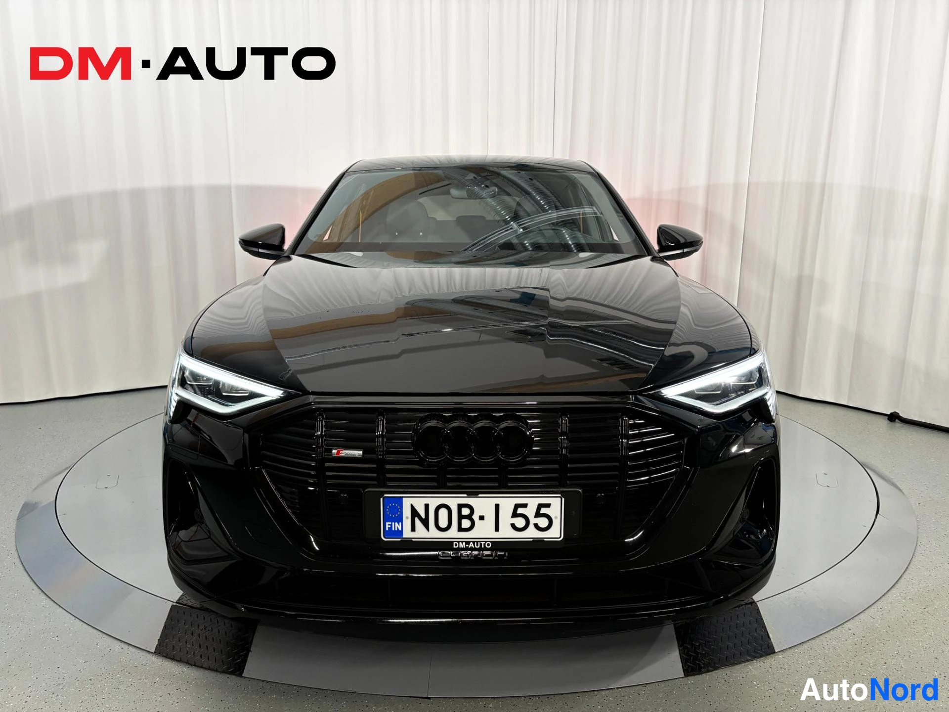Edestä: Mustan Audi Sportbackin suora etunäkymä: suuri etusäleikkö, S-merkintä ja rekisteritunnus, auton ympärillä valkoinen verho.