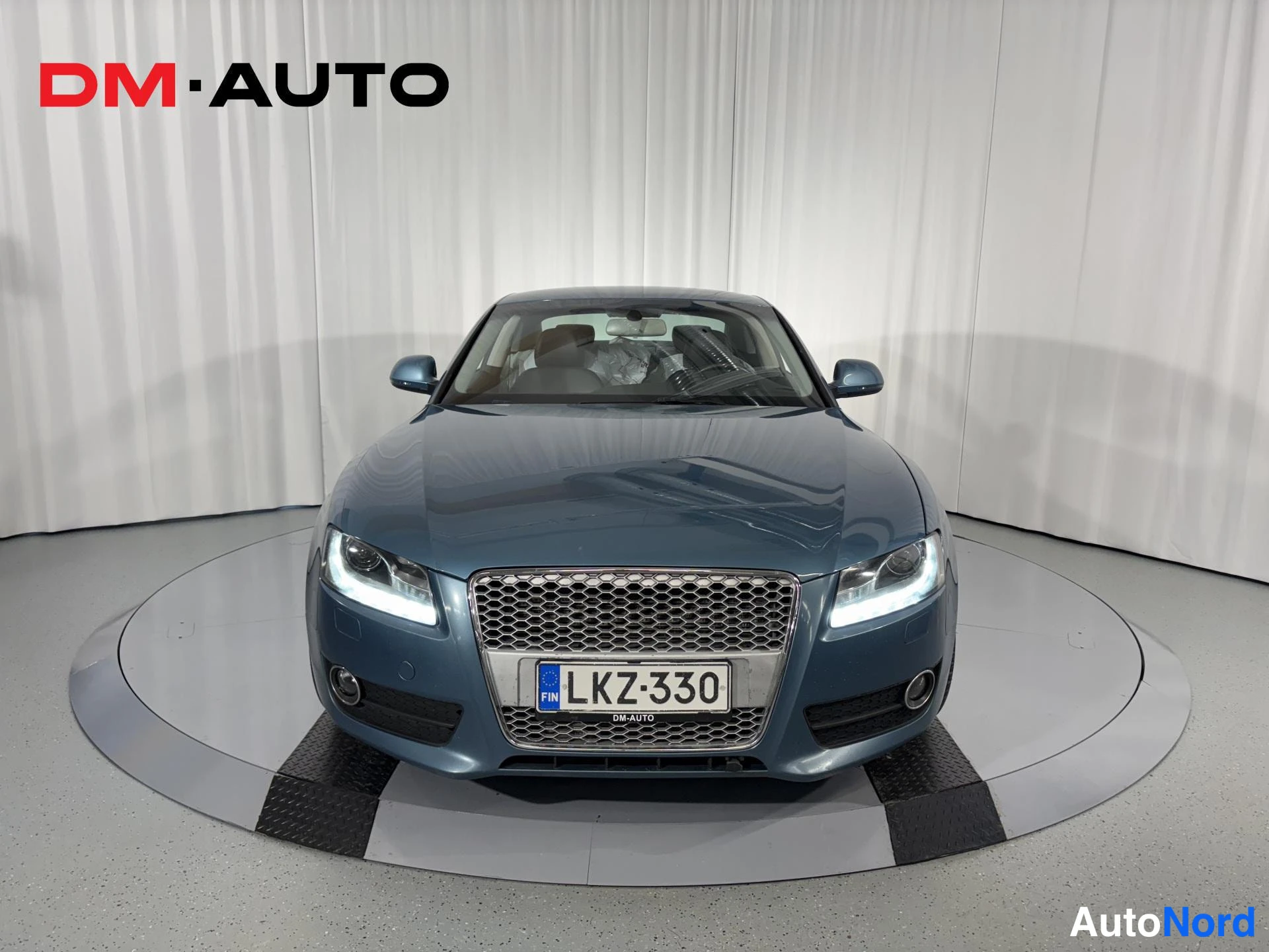 Edestä: Sinisenharmaa Audi A5 suoraan edestä, etusäleikkö ja rekisterikilpi näkyvissä.