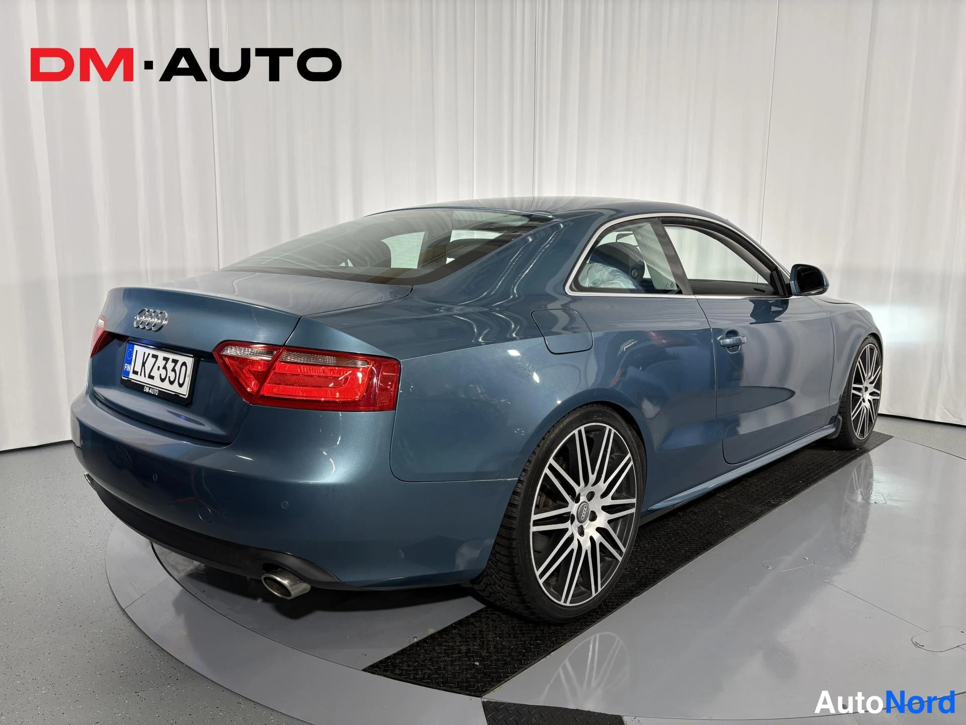 Takaviisto: Sininen Audi A5 -coupe kulmassa takaa kuvattuna, näkyvissä takavalo, rekisterikilpi ja hopeamonet vanteet.