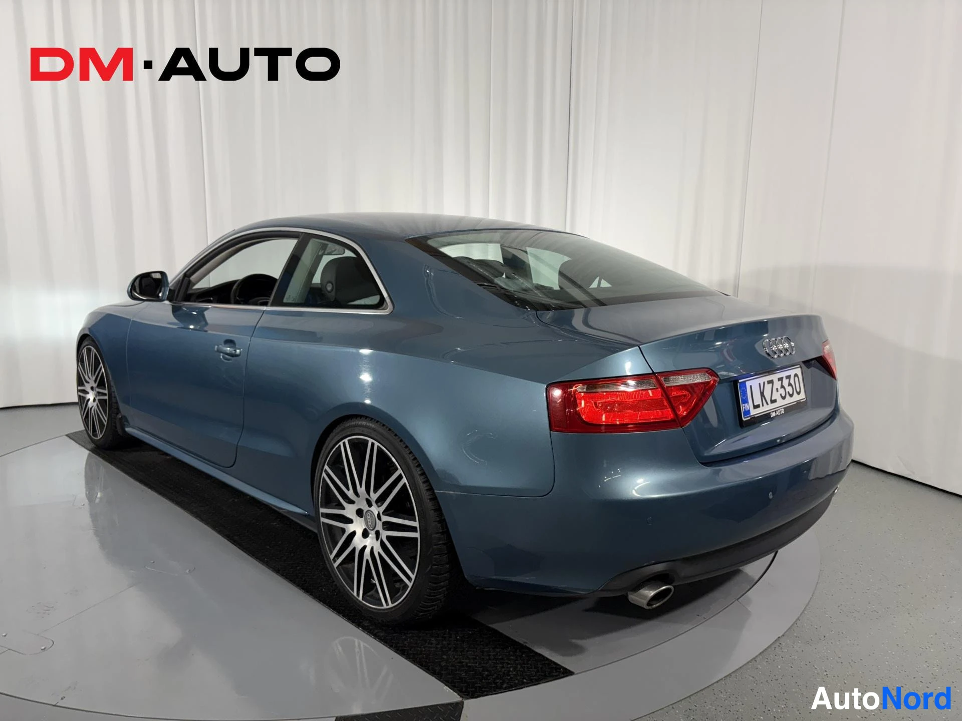 Takaviisto: Sininen Audi A5 -coupe kulmassa takaa vasemmalta kuvattuna, näkyvissä takavalo, rekisterikilpi ja hopeamonet vanteet.