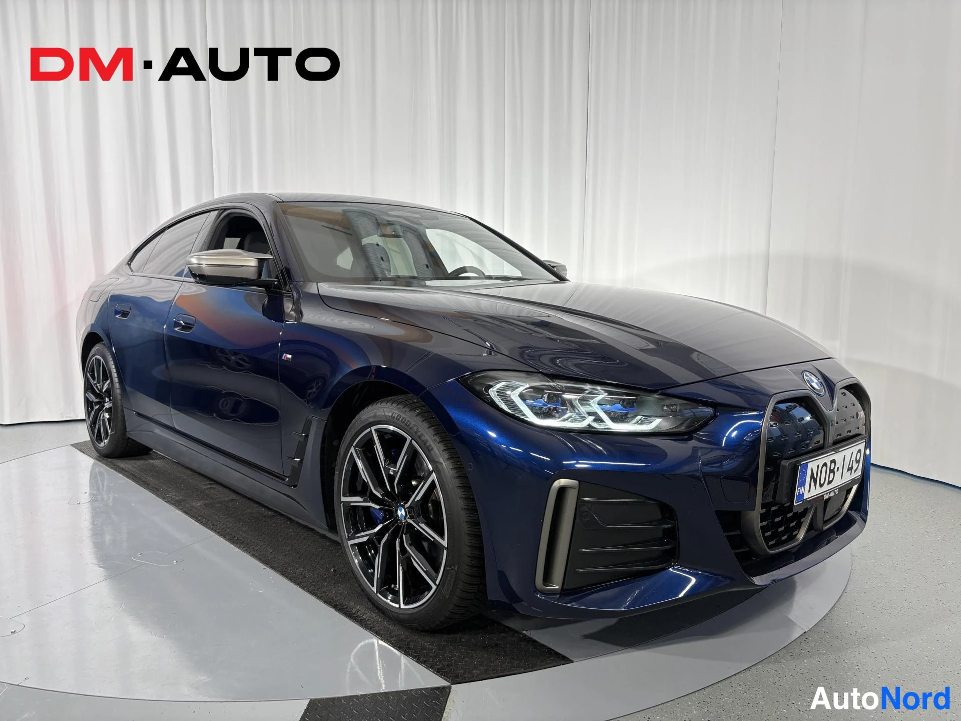 Etuviisto, nokka oikealle: Tummansininen BMW i4 kulmassa edestä oikealta kuvattuna, etusäleikkö, ajovalot ja mustat kevytmetallivanteet näkyvät.