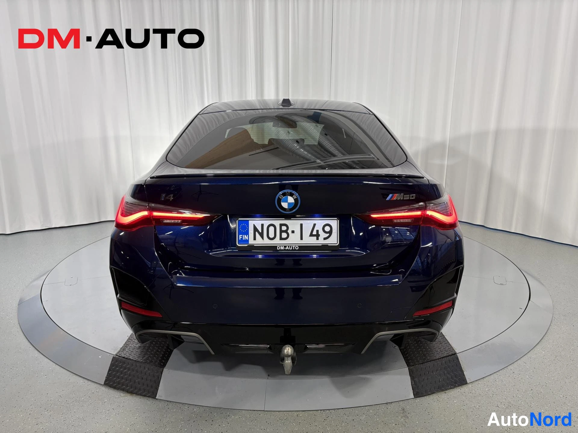 Takaa: BMW:n suora takaosakuva: tummansininen korirakenni, i4- ja M50-merkinnät, N0B-149-kilpi ja perävaunukoukun pallo näkyvät.