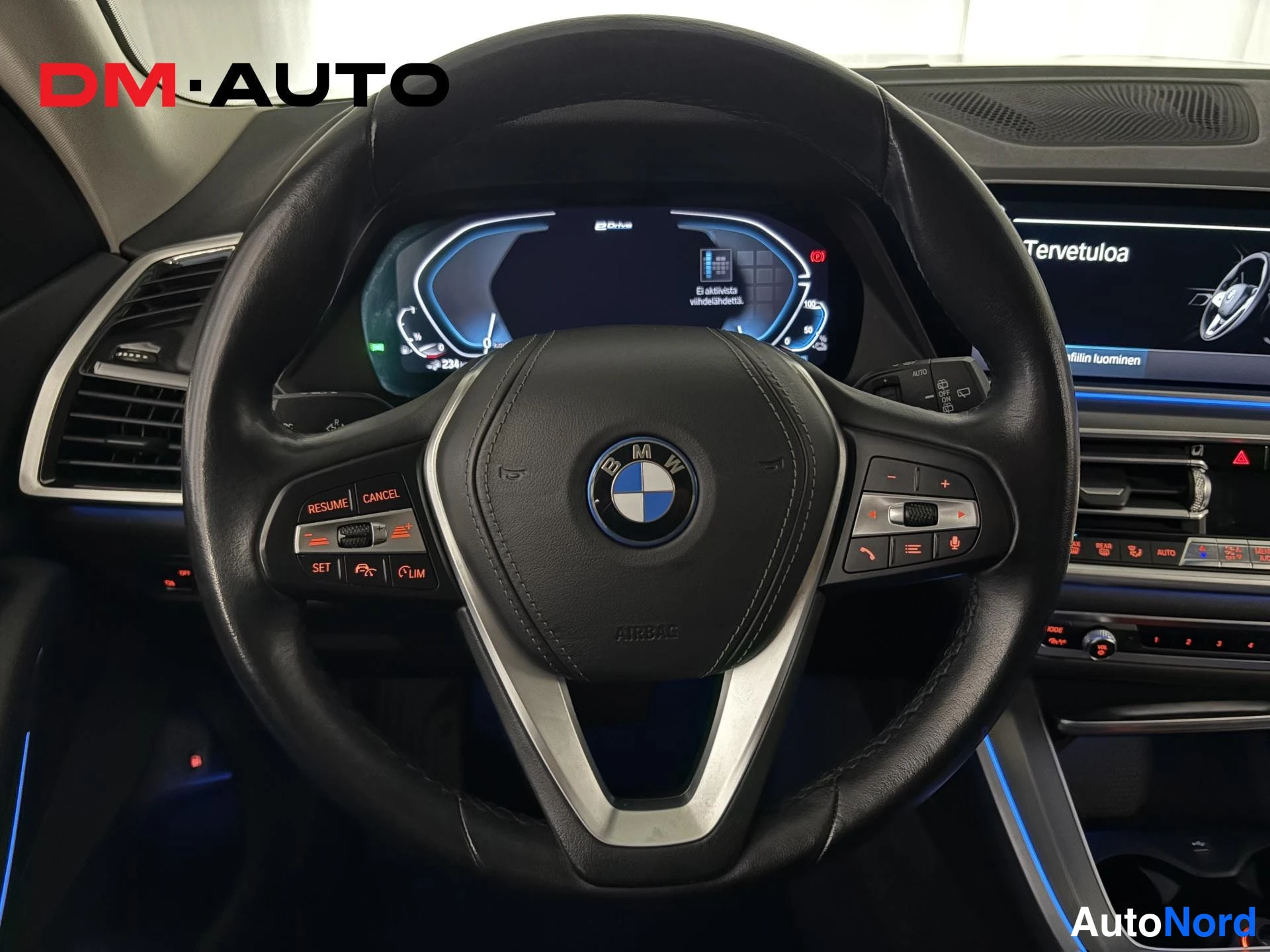 Muu: Lähikuva BMW:n ohjauspyörästä ja digitaalisesta mittaristosta; keskuskonsolin näytöllä lukee 'Tervetuloa'.