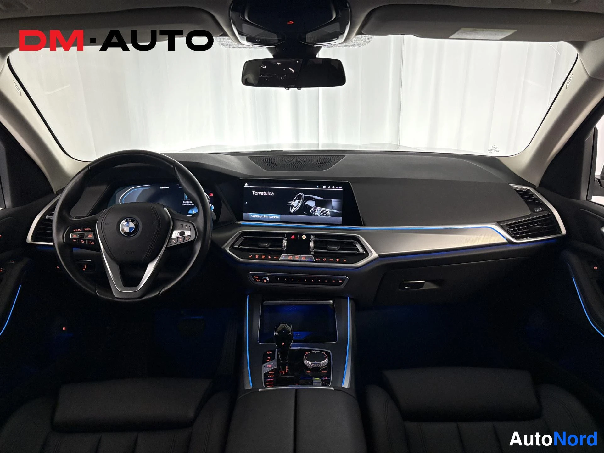 Muu: BMW:n ohjaamosta eteenpäin kuvattu näkymä: ohjauspyörä, tietoinfotainment-näytöllä teksti 'Tervetuloa', keskikonsoli ja tummat penkit.