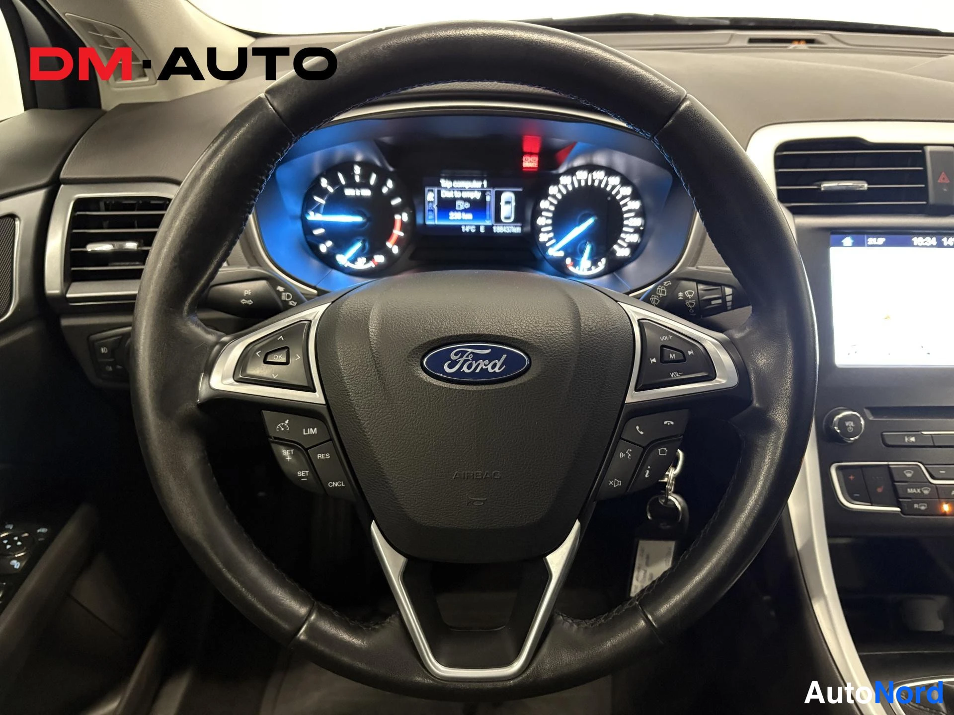 Muu: Ford-ohjauspyörän lähikuva Ford Mondeon sisällä, taustalla kaareva mittaristo ja osa infotainment-näytöstä; ruudulla näkyy Ilmastointi-valikko.