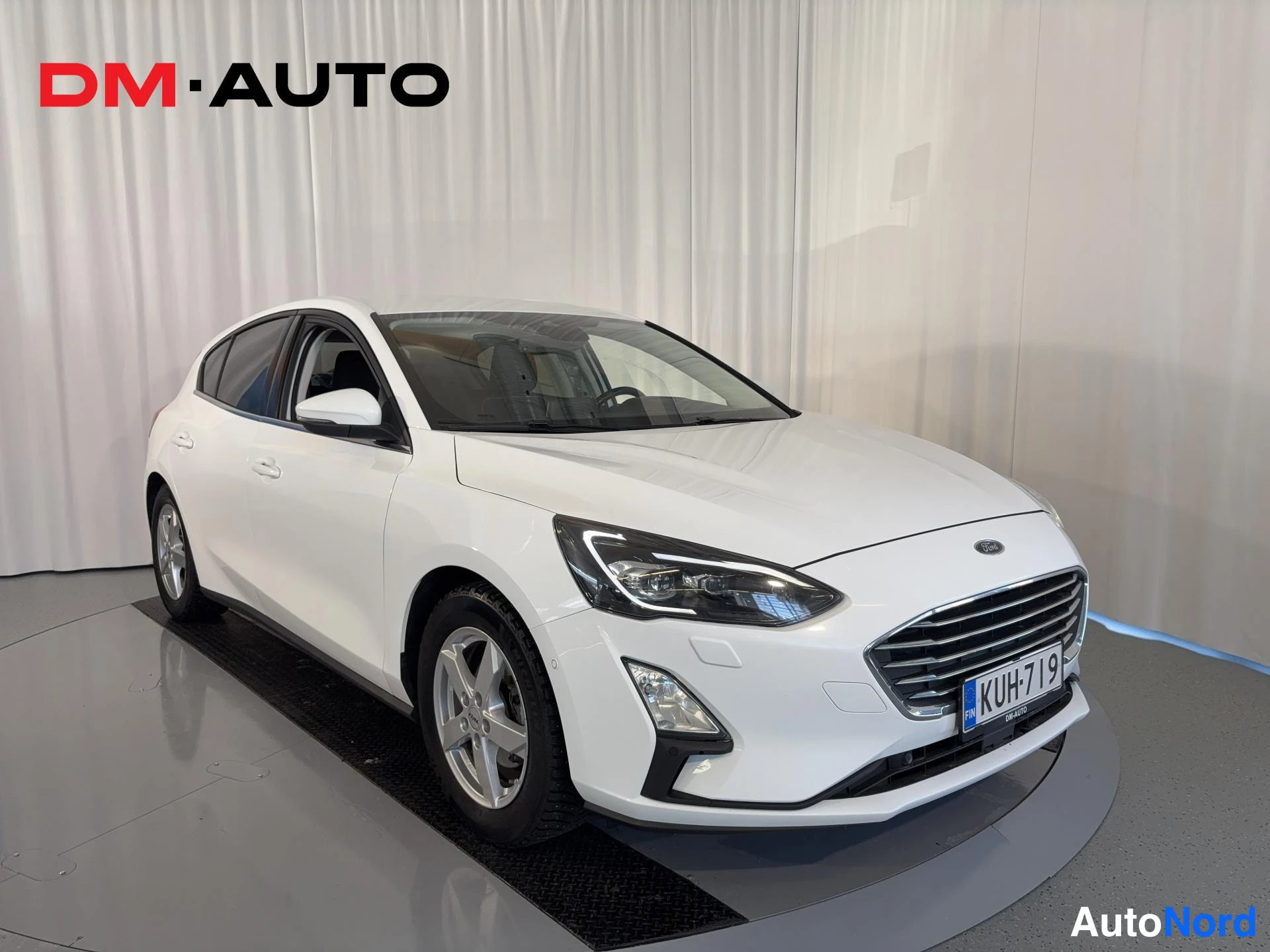 Etuviisto, nokka oikealle: Valkoinen Ford Focus -henkilöauto etu kolmasosakulmasta, keula oikealla, rekisterikilpi.