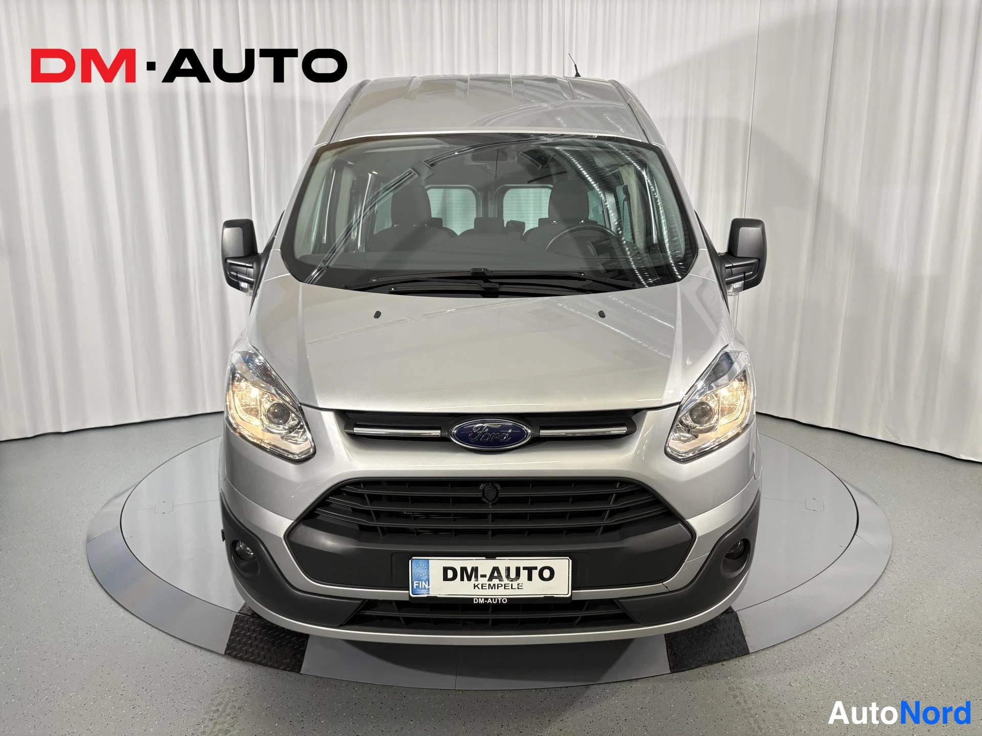 Edestä: Hopeanharmaan Ford Transit Custom -pakettiauton etunäkymä, jossa näkyvät etugrilli, Ford-merkki ja ajovalot.