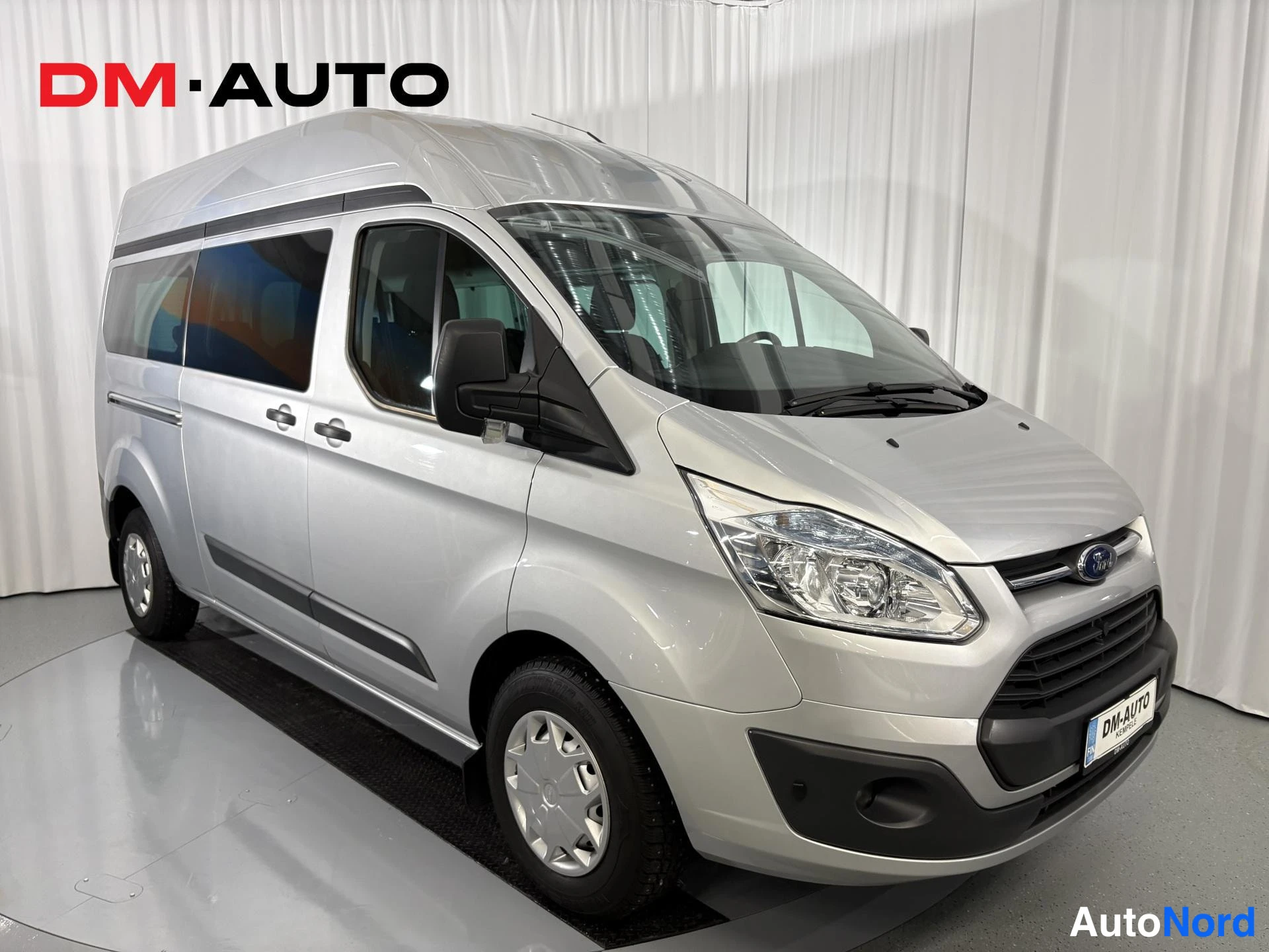 Etuviisto, nokka oikealle: Hopeanharmaa Ford Transit Custom -pakettiauto etuosalta ja oikealta sivulta kuvattuna studiossa.