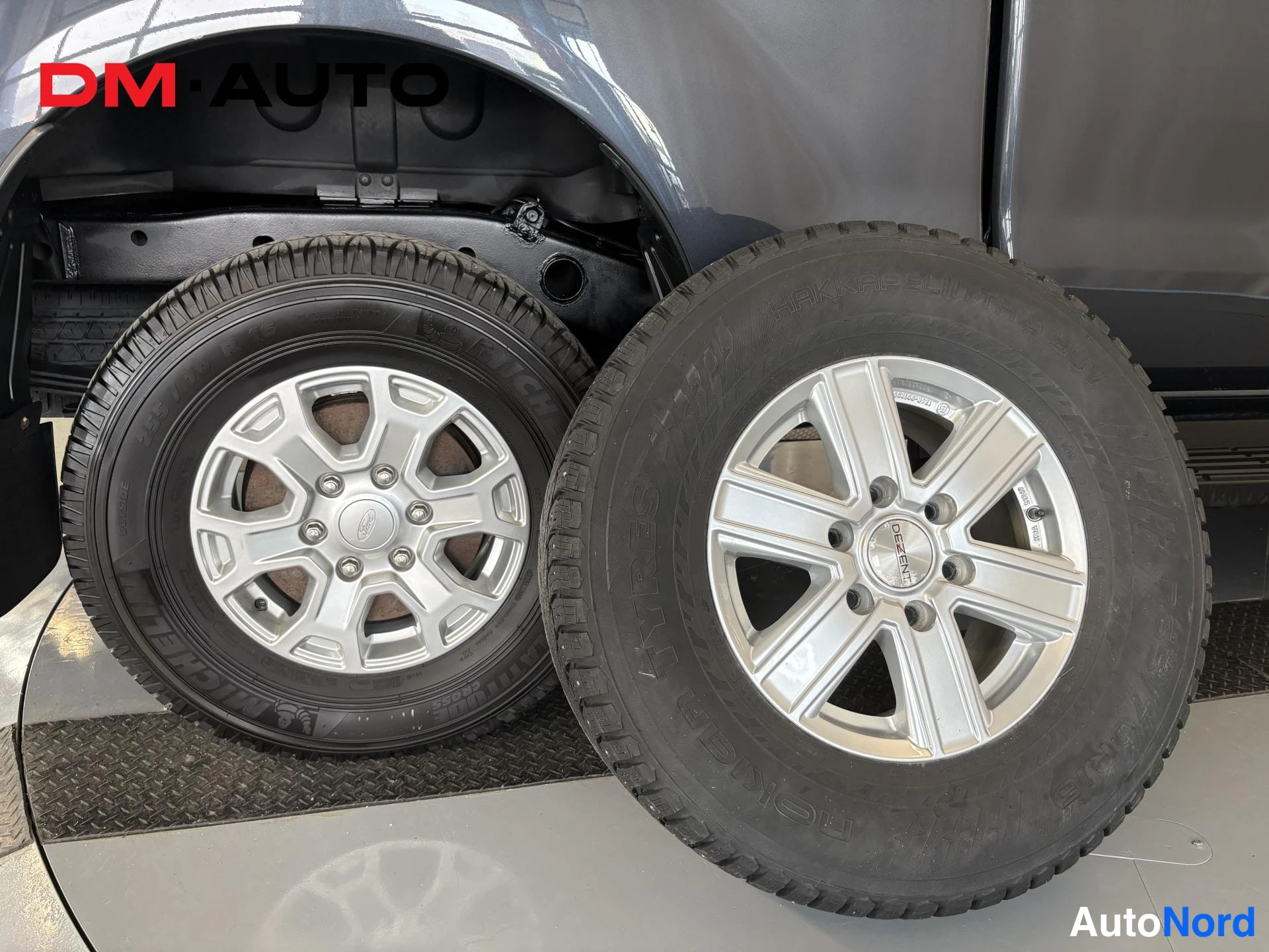 Muu: Kaksi vanteellista rengastta: autosta irrotettu Ford-merkitty hopea vannas ja sen vieressä toinen hopea vannas niititetyllä nailgummusta rengasta, musta koripel