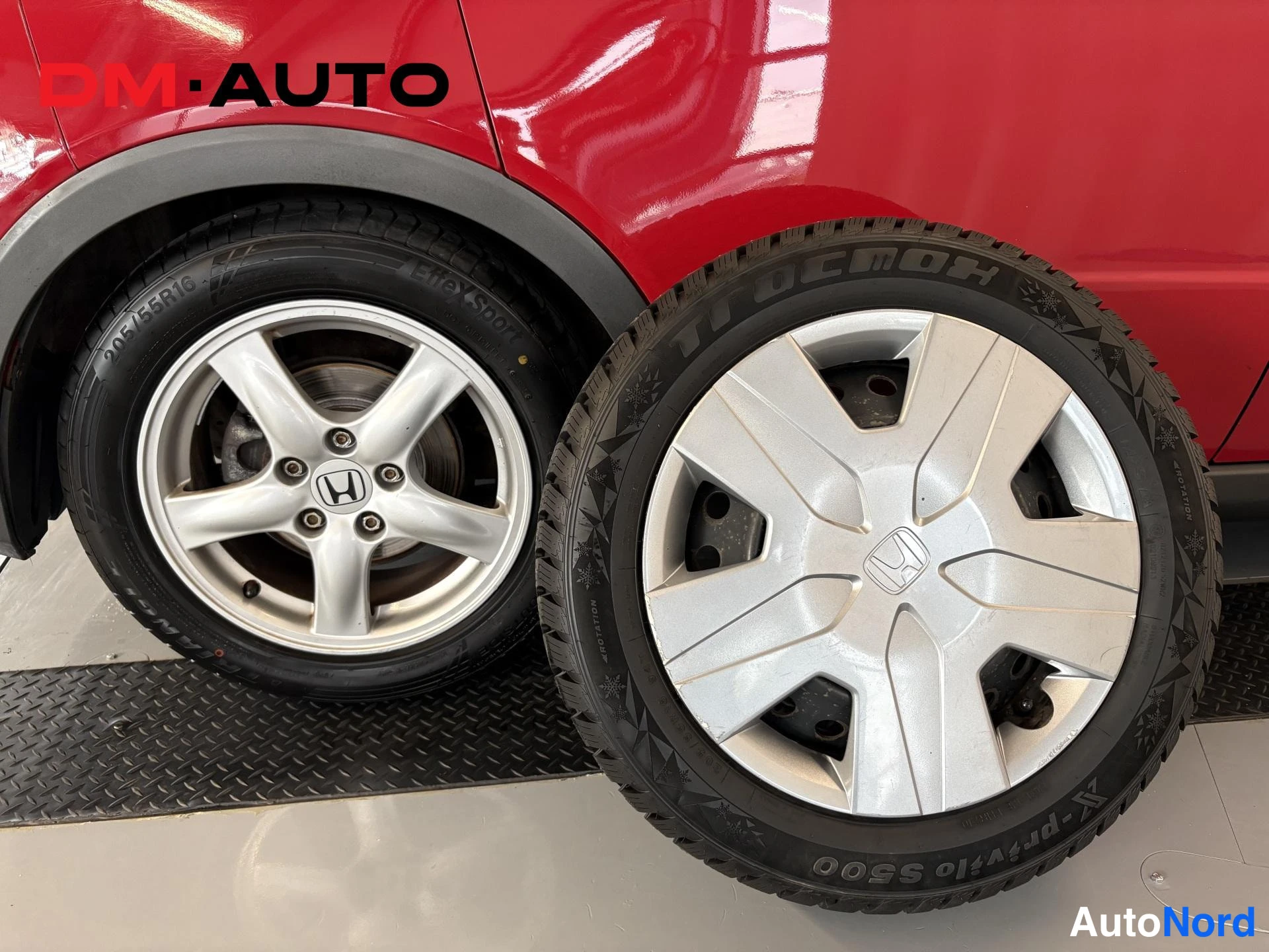 Etuviisto, nokka oikealle: Punaisen auton hopea kevytmetallivanne (205/55 R16) ja vieressä alla oleva nailonkitkarenkaittaen Hakka Blue -pintainen varapyörä Honda-merkkisellä pölyk