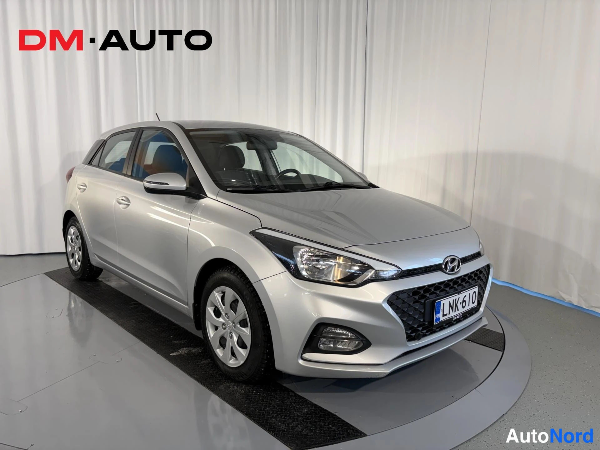 Etuviisto, nokka oikealle: Hopeanvärinen Hyundai i20 etukulmasta oikealta, etusäleikkö ja ajovalot näkyvissä, rekisterikilpi.