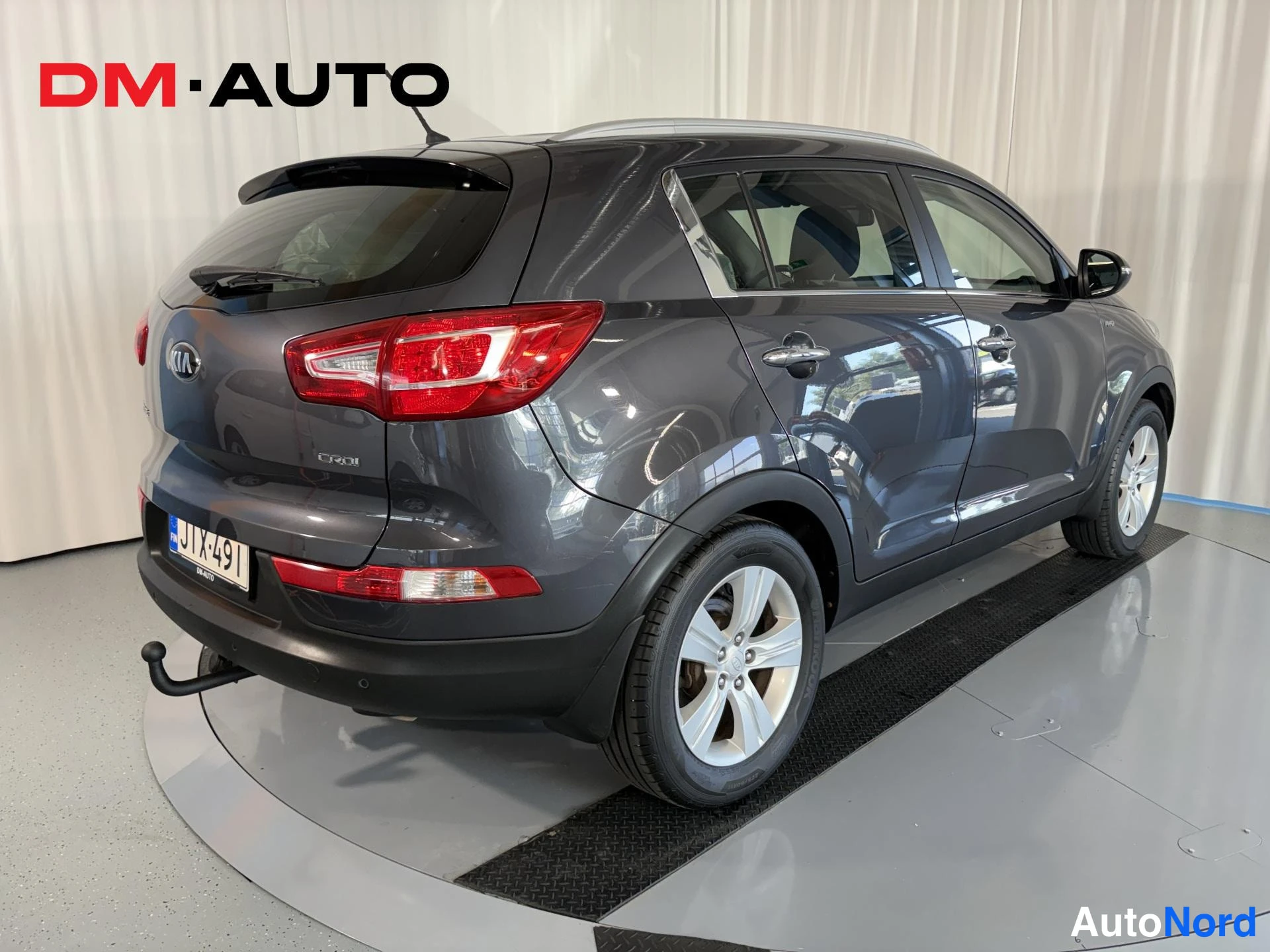 Takaviisto: Auton takaosa ja vasen sivu kolmen neljänneskulmasta: tummanharmaa Kia Sportage, punaiset takavalot, perävaunukoukun kaula ja rekisterikilpi näkyvät.
