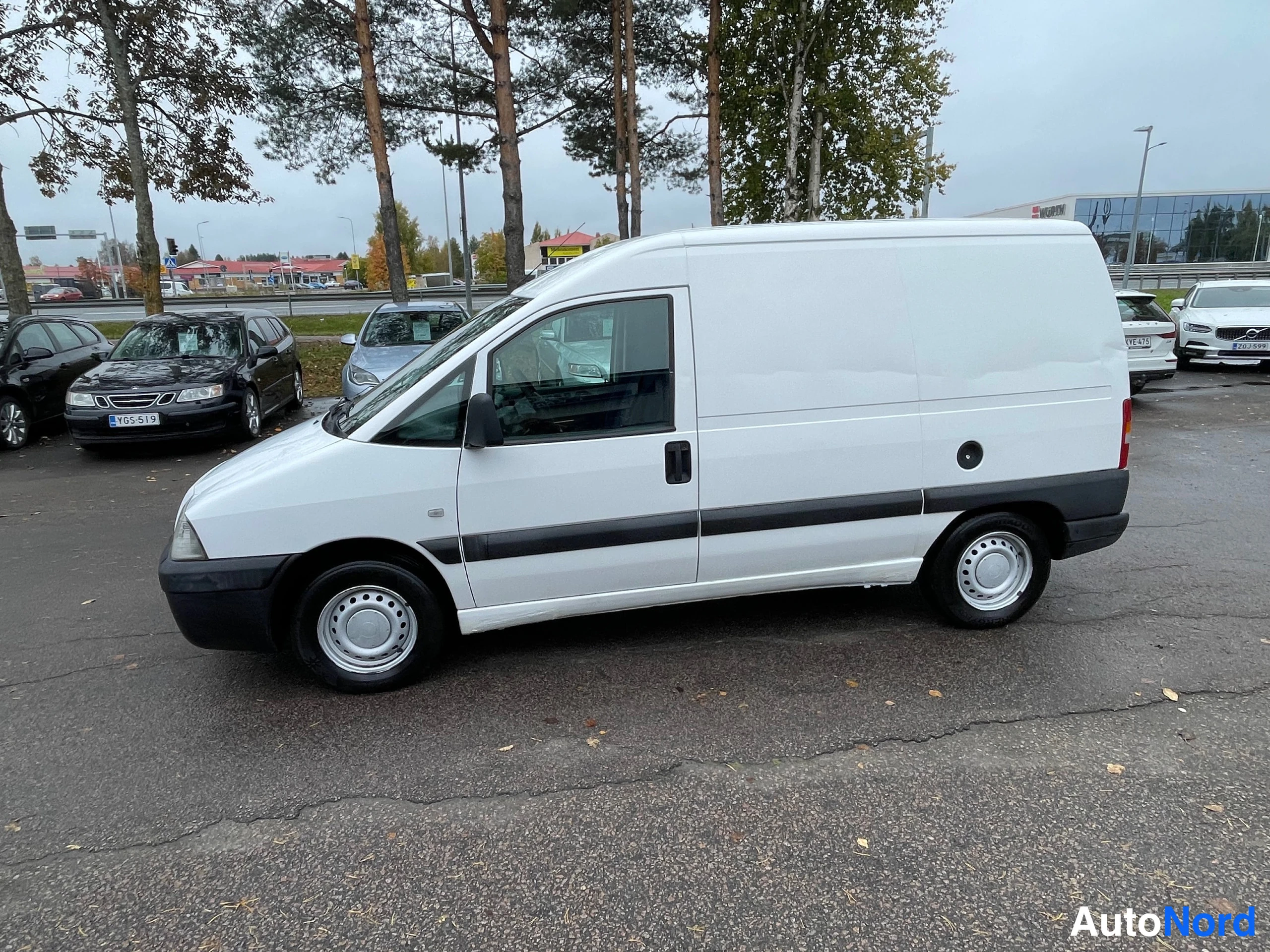 Etuviisto, nokka vasemmalle: Valkoinen Fiat Scudo -pakettiauto sivulta etukulmasta, keula vasemmalla, autopaikalla muitten autojen joukossa.