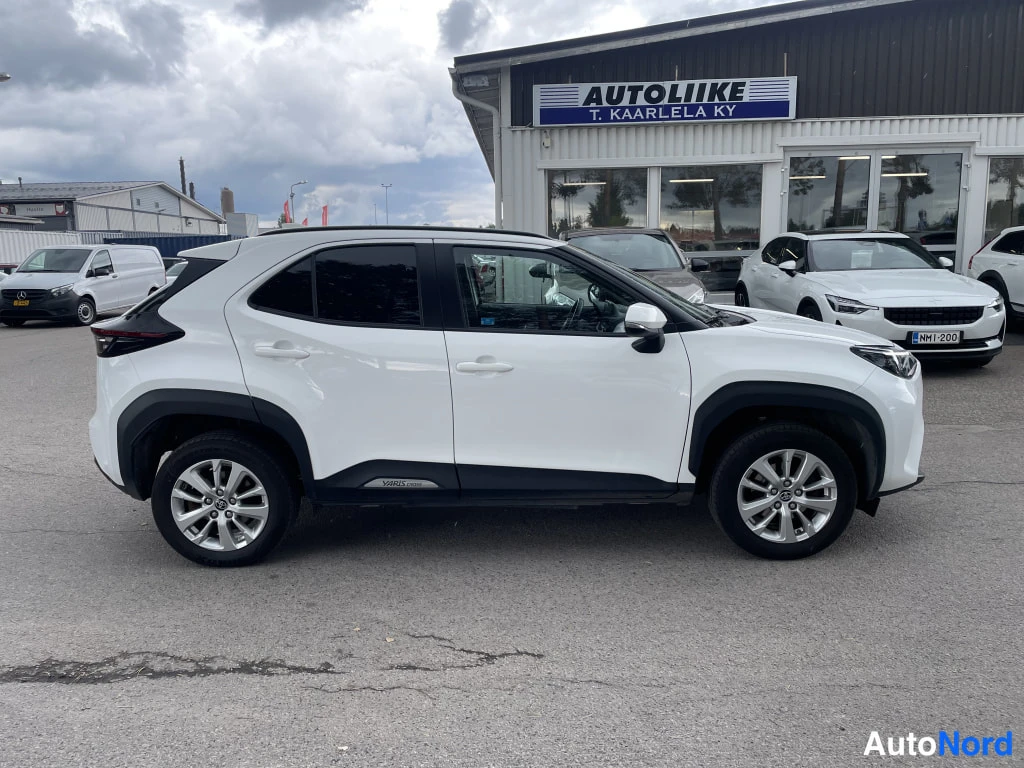 Etuviisto, nokka oikealle: Valkoinen Toyota Yaris Cross sivulta etukulmasta, keulaa oikealla, autoliikkeen pihalla.