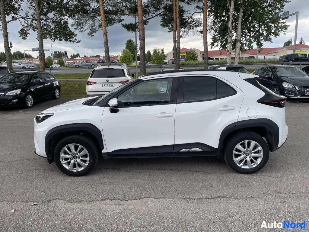 Etuviisto, nokka vasemmalle: Valkoinen Toyota Yaris Cross sivulta etukulmasta, keulaa vasemmalla, pysäköintialueella muiden autojen joukossa.