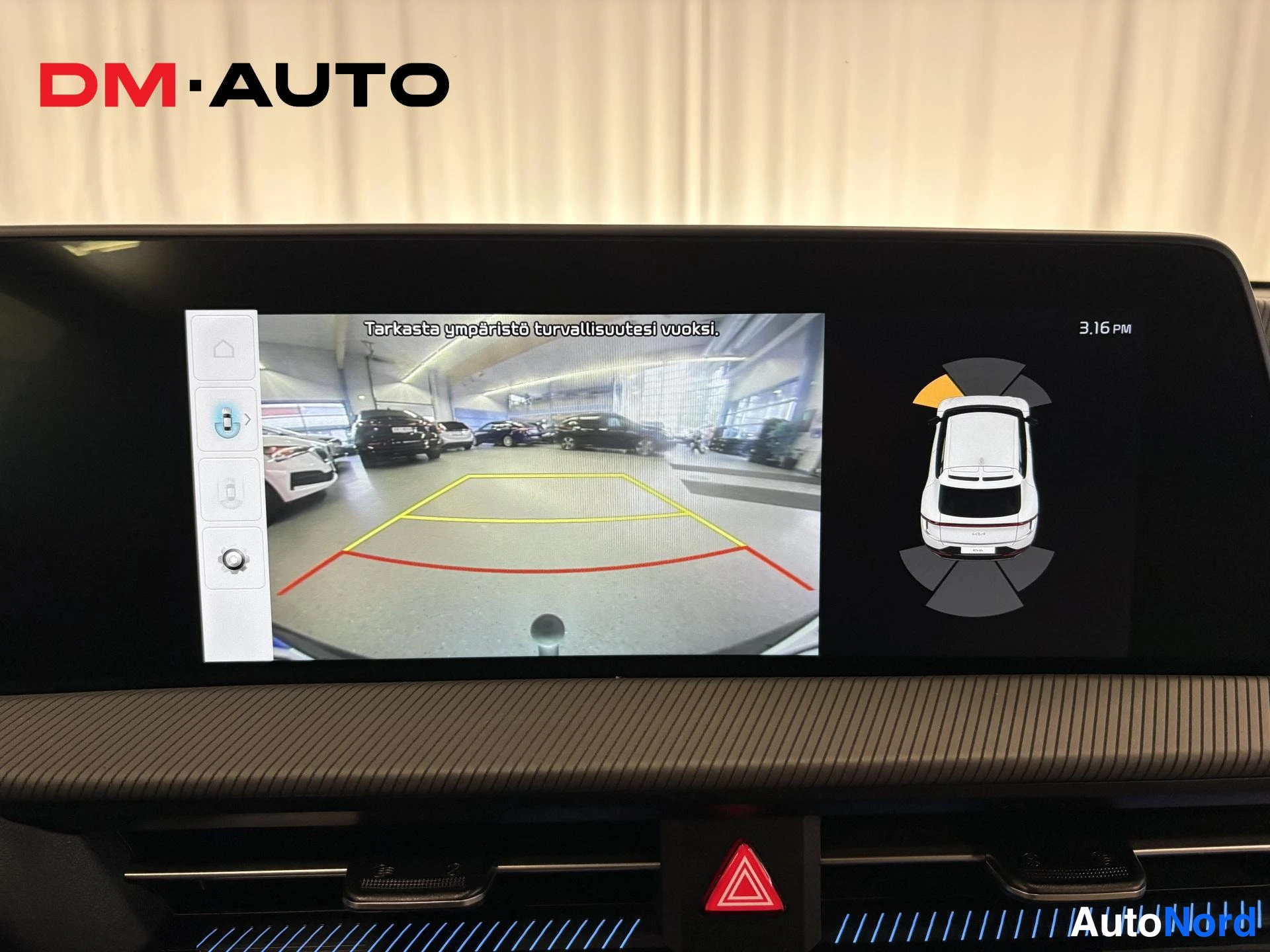 Muu: Infotainment-näytöllä näkyy peruutuskameran kuva hyppyriviin ja ympäristönäyttö autojen reunaviivatyyppisin ohjeviivoin, teksti 'Tarkasta ympäristö turvallisuut