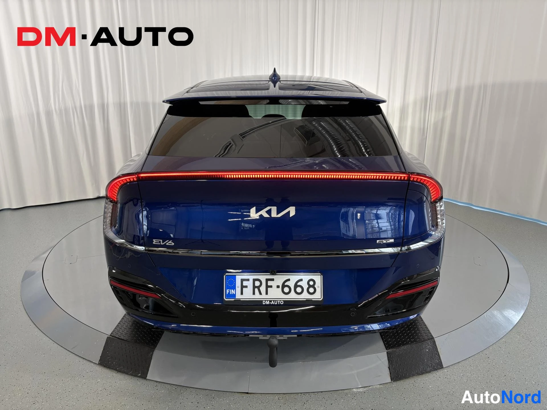 Takaa: Sininen Kia EV6 suoraan takaa; näkyvissä KIA-merkki, EV6-teksti, kokonaan poikki kulkeva punainen takavalojuova, -rekisterikilpi ja perävaunukoukku.
