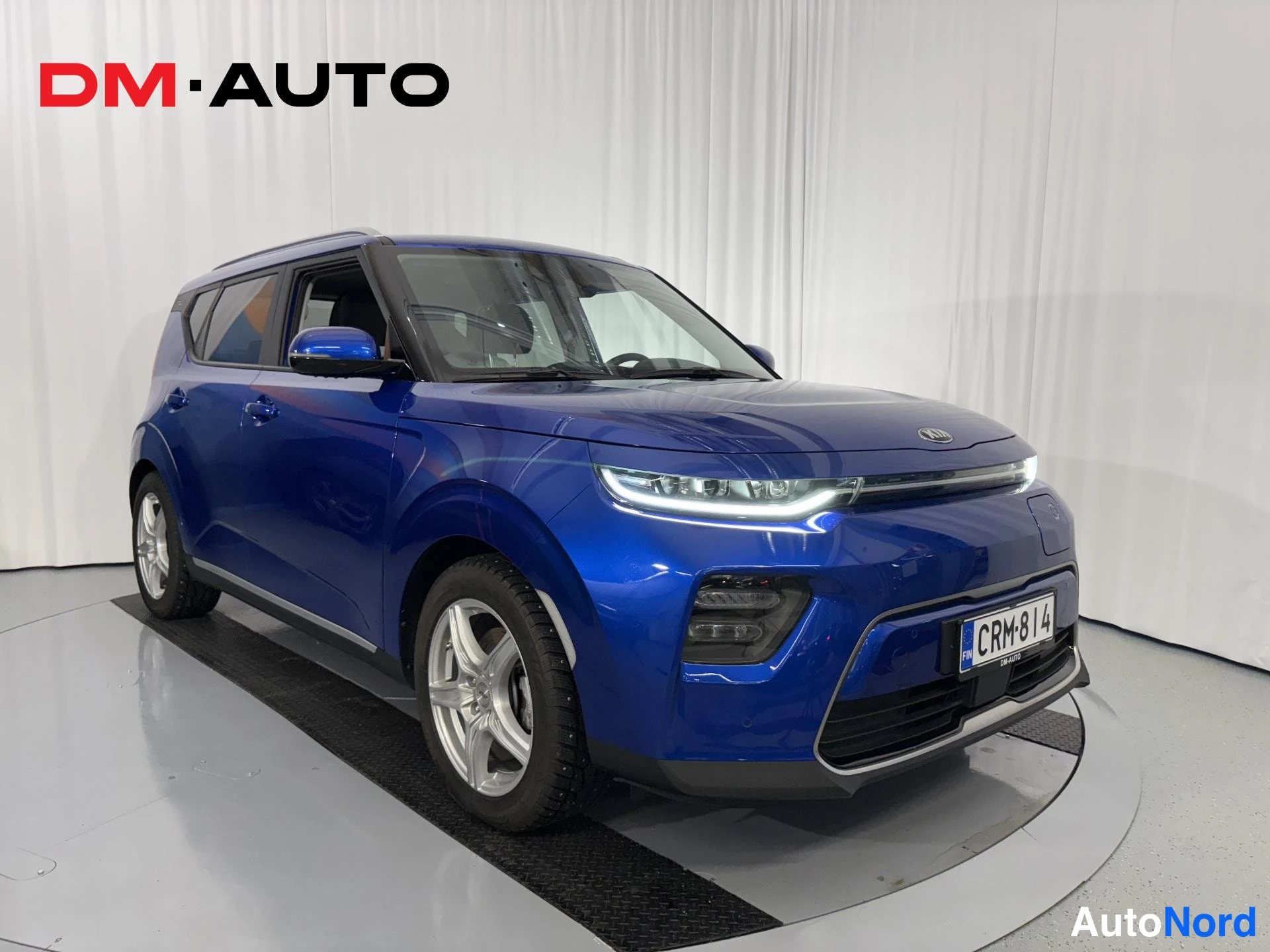 Etuviisto, nokka oikealle: Sininen Kia e-Soul etuviistosta oikealta, etumoitto ja etupuskuri sekä oikea kylki valkaisualustalla.