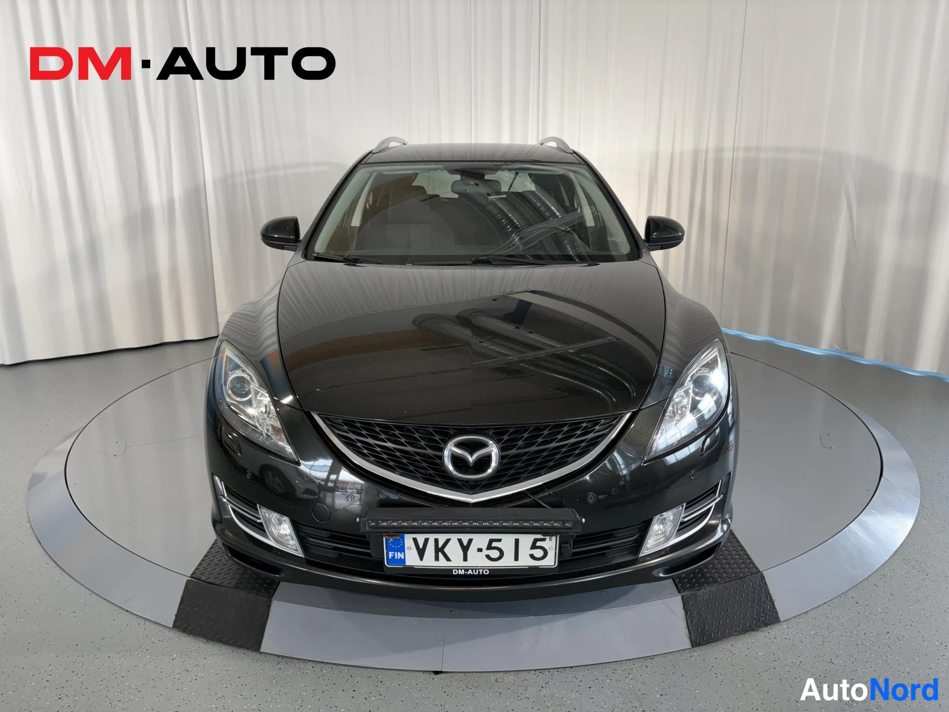 Edestä: Mustan Mazda 6 farmarin suora etunäkymä, Mazda-merkki keulassa ja rekisterikilpi, DM-AUTO-logon alla.