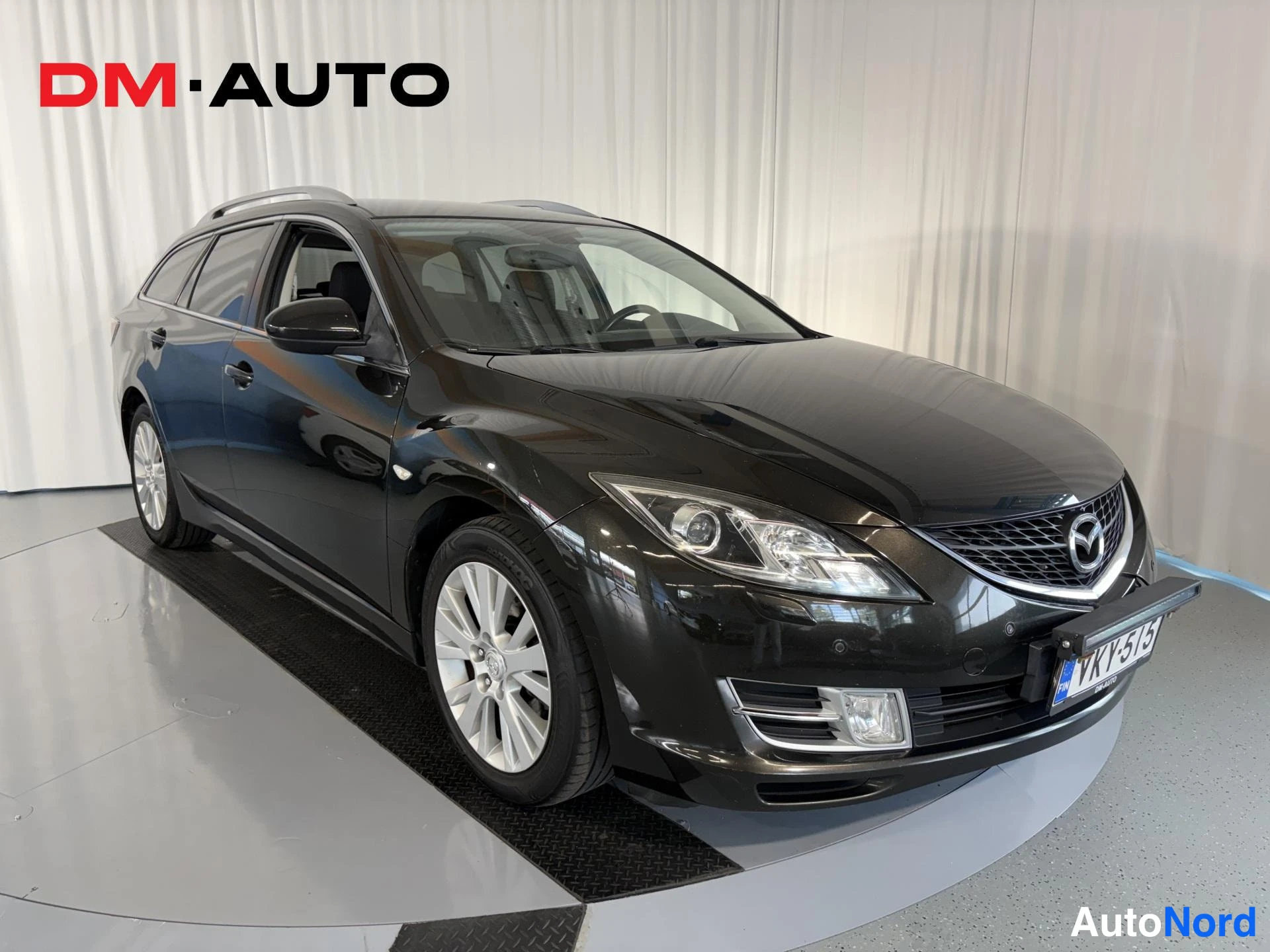 Etuviisto, nokka oikealle: Musta Mazda 6 farmari etukulmasta oikealta kuvattuna, keula ja kylki näkyvissä, rekisterikilpi keulassa.