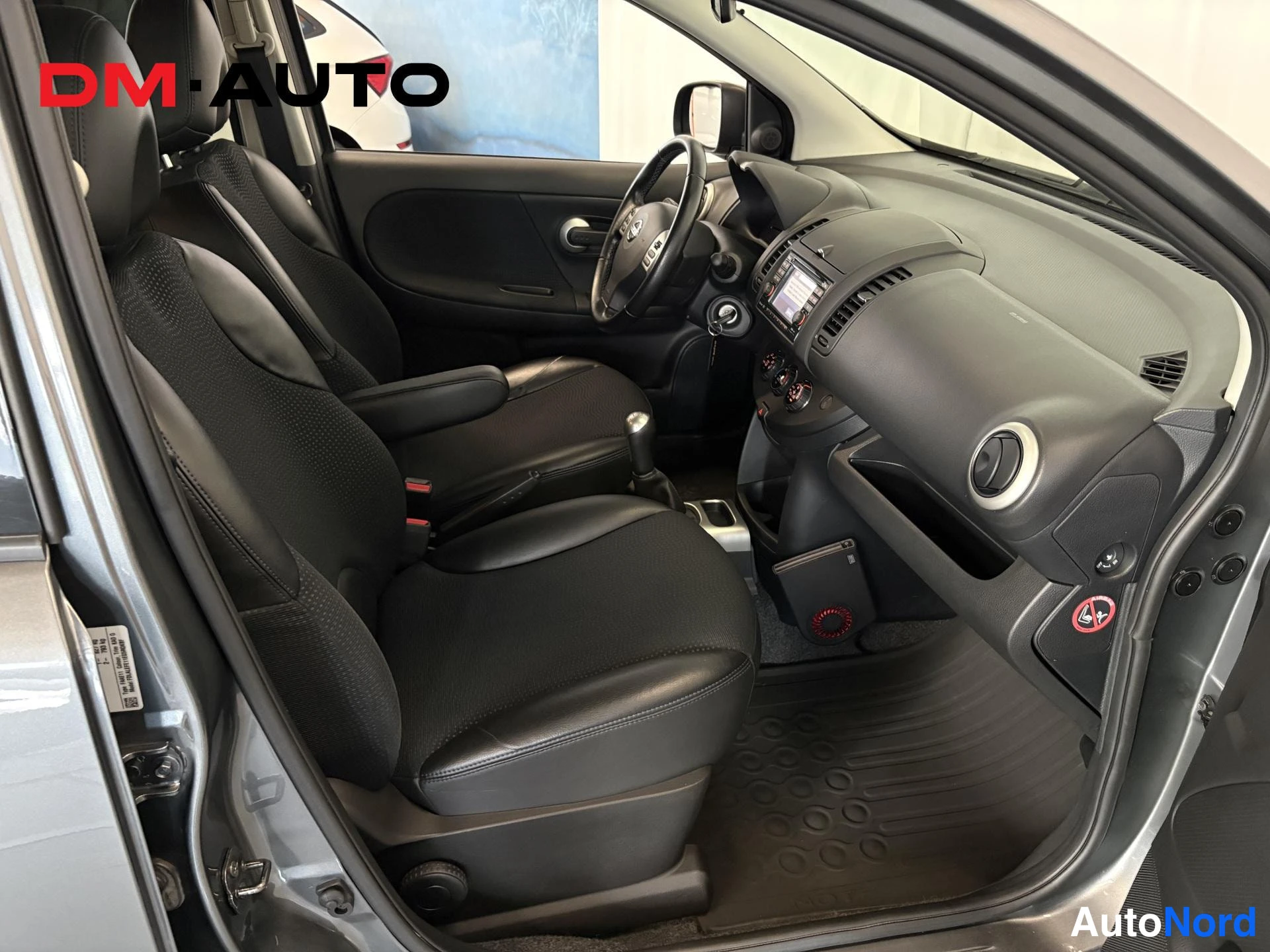 Muu: Nissan Note:n etusisätilat kuljettajan ovesta katsottuna: ohjauspyörä, kojelauta, infotainment-näyttö ja manuaalivaihteenKeihä on näkyvissä; penkit mustaa nahka