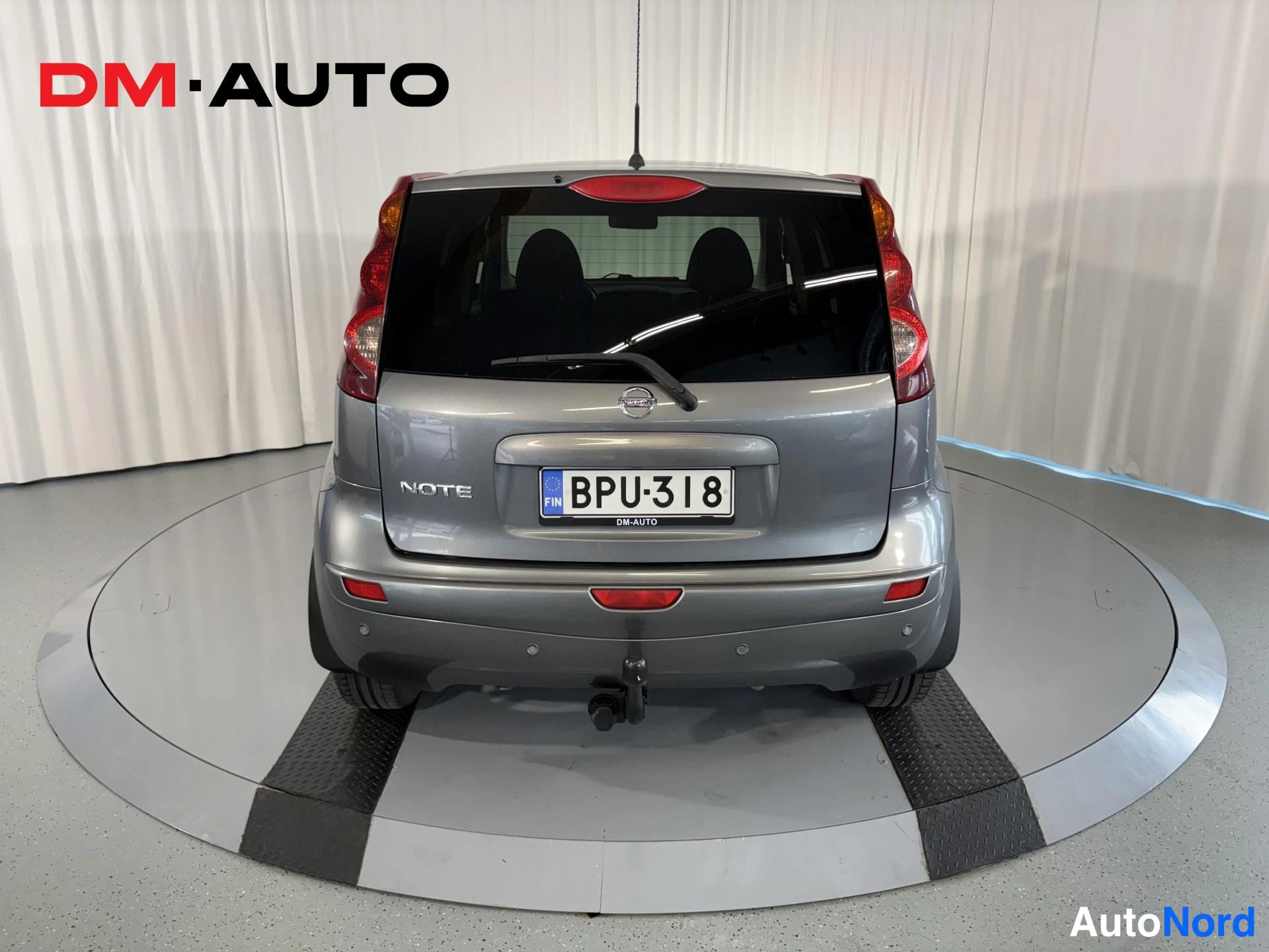 Takaa: Auton takaosa suoraan takaa: harmaa Nissan Note -takapaneeli, takaluukun lasi, takavalot, rekisterikilpi ja perävaunukytkin.