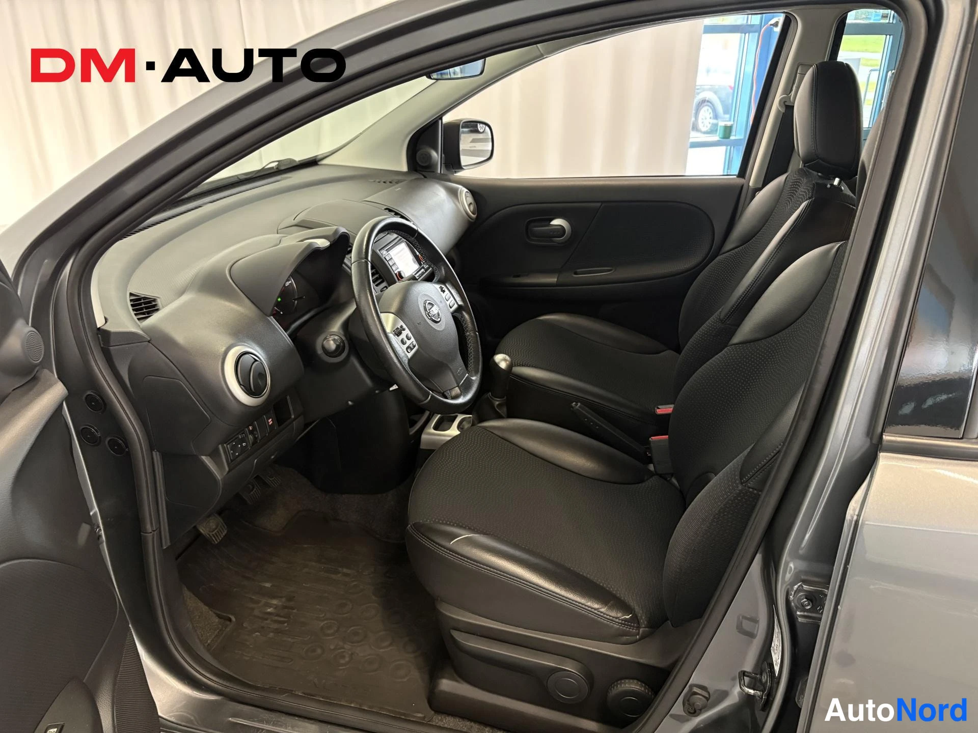 Muu: Avoimesta kuljettajan ovesta kuvattu Nissan Note:n sisätilat: tummat etuistuimet, ohjauspyörä, kojelauta ja manuaalivaihtoheidin.