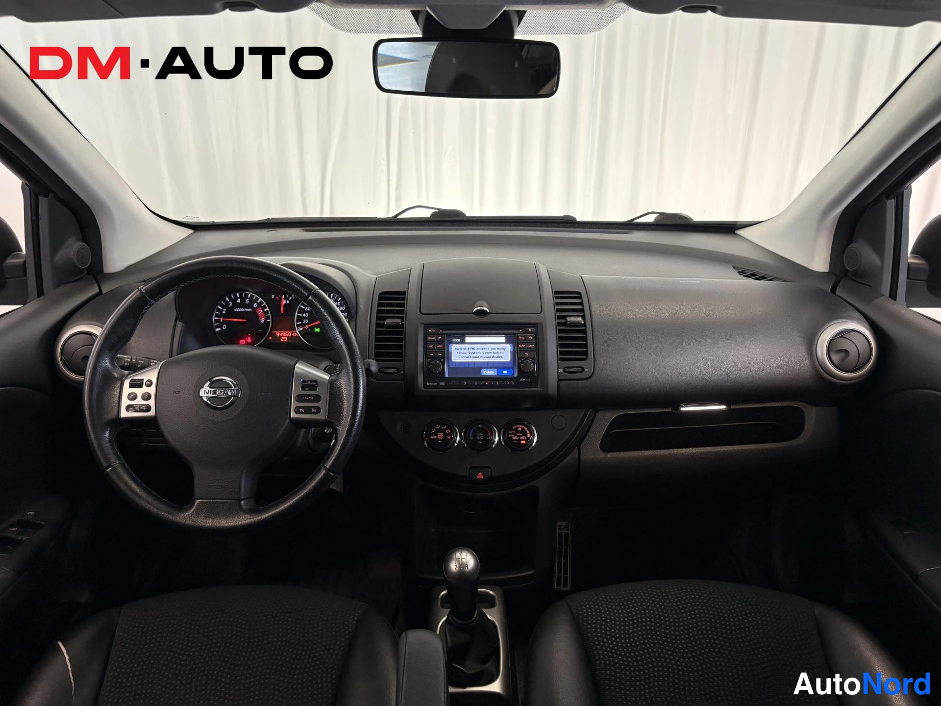 Muu: Näkymä Nissan Note:n kojelautaan tuulilasin läpi: ohjauspyörä, mittaristo, keskikonsolen radionäyttö ja kolme säätönuppia, manuaalivaihtoheinä.