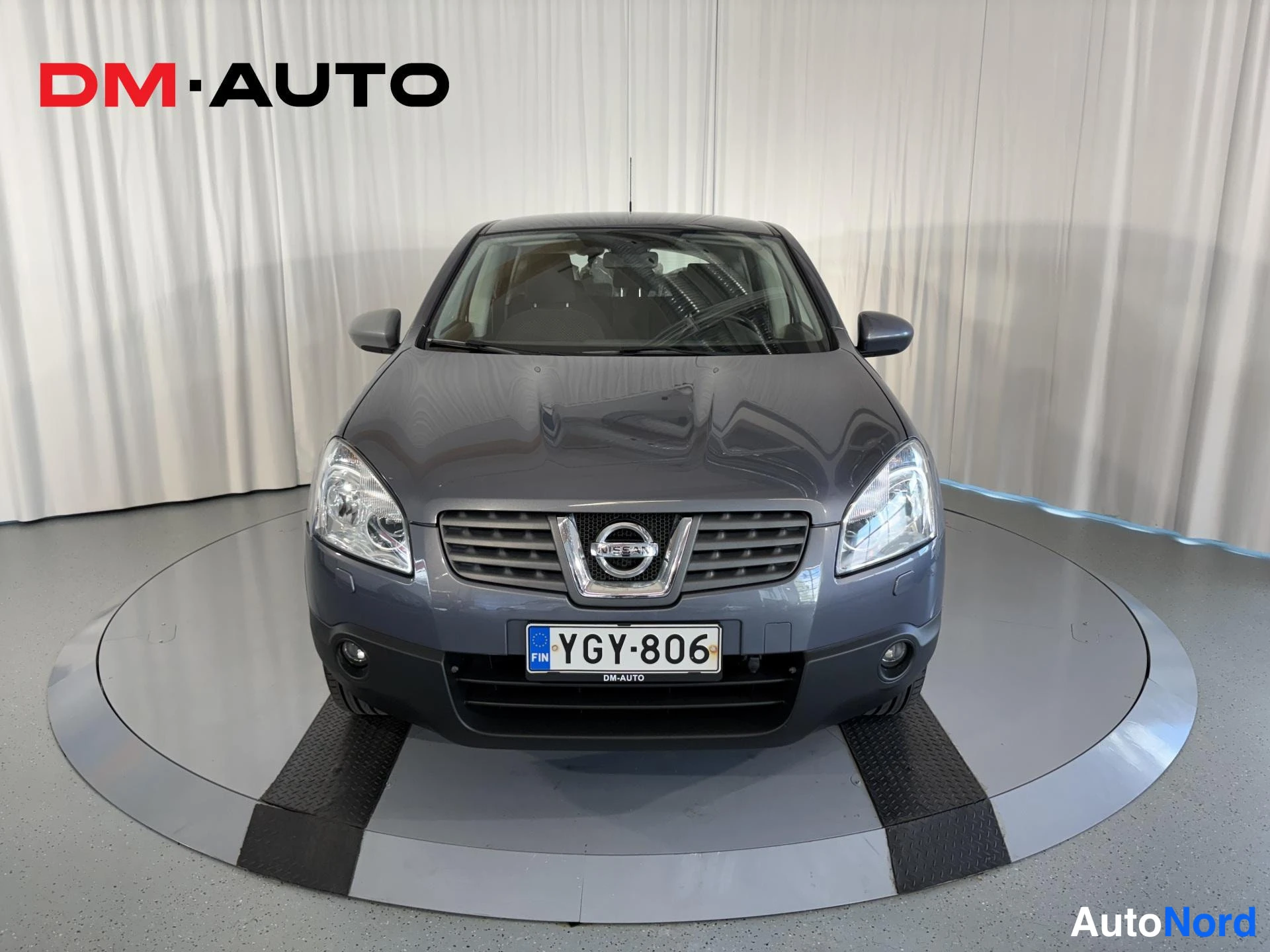 Edestä: Nissan Qashqai, harmaa, suora etänäkymä studioissa, rekisterikilpi ja DM-AUTO-teksti kilven alla.