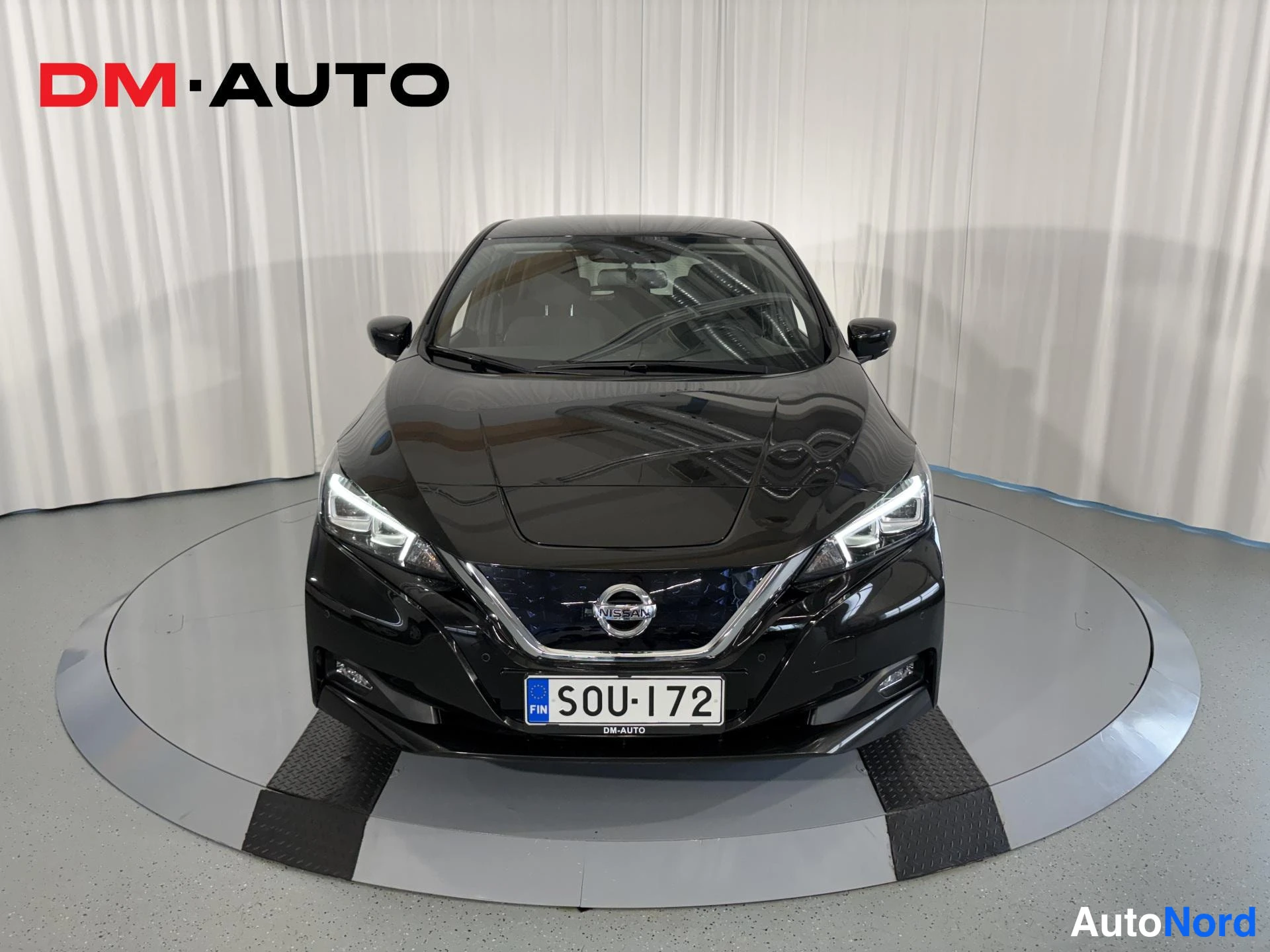 Edestä: Mustan Nissan Leafin etupuoli suoraan edestä, keulavalo paistaa, rekisterikilpi.
