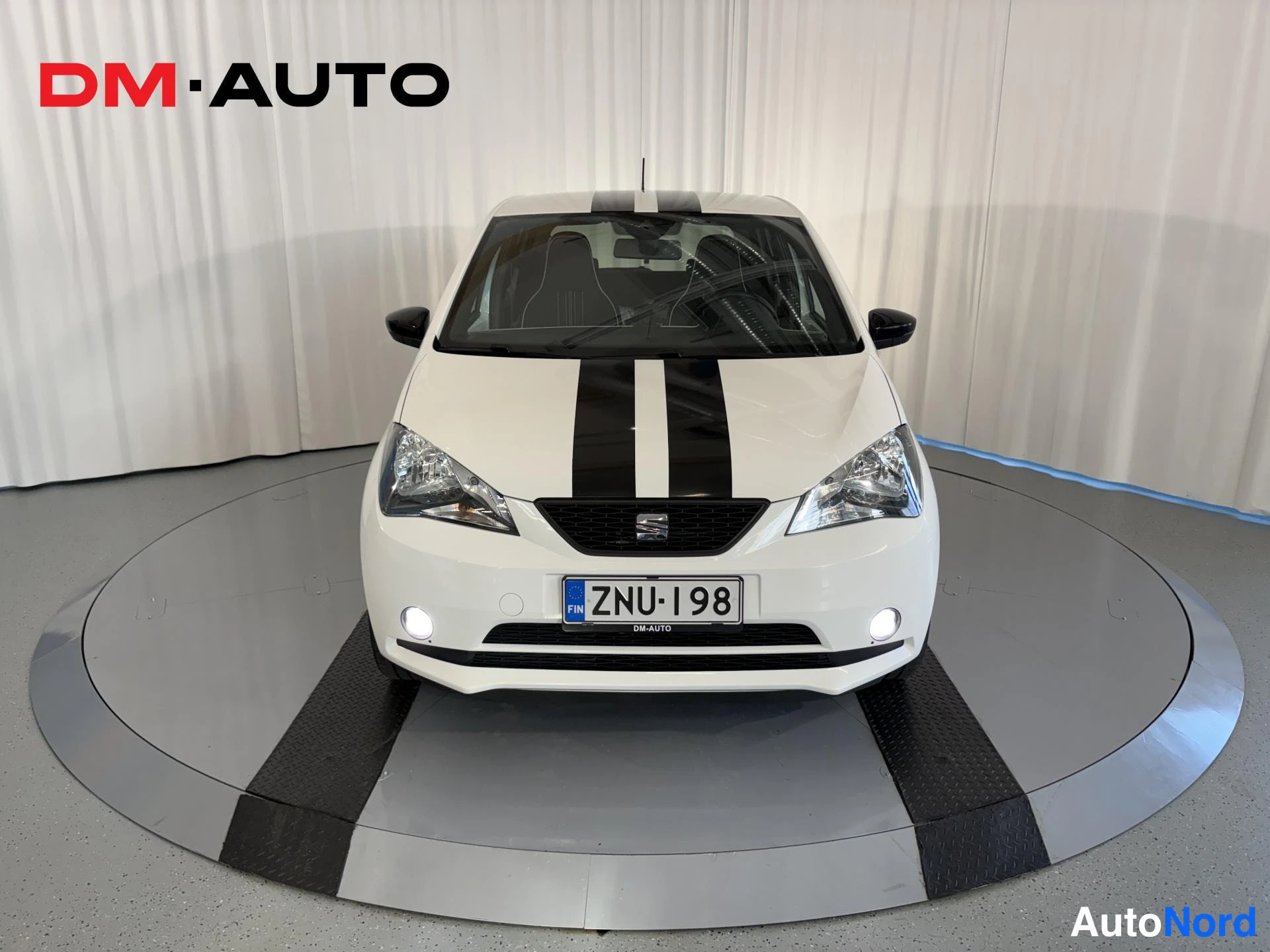 Edestä: White SEAT Mii Electric photographed directly from the front: grille, headlights, front bumper and number plate are visible, with a black double stripe on the b