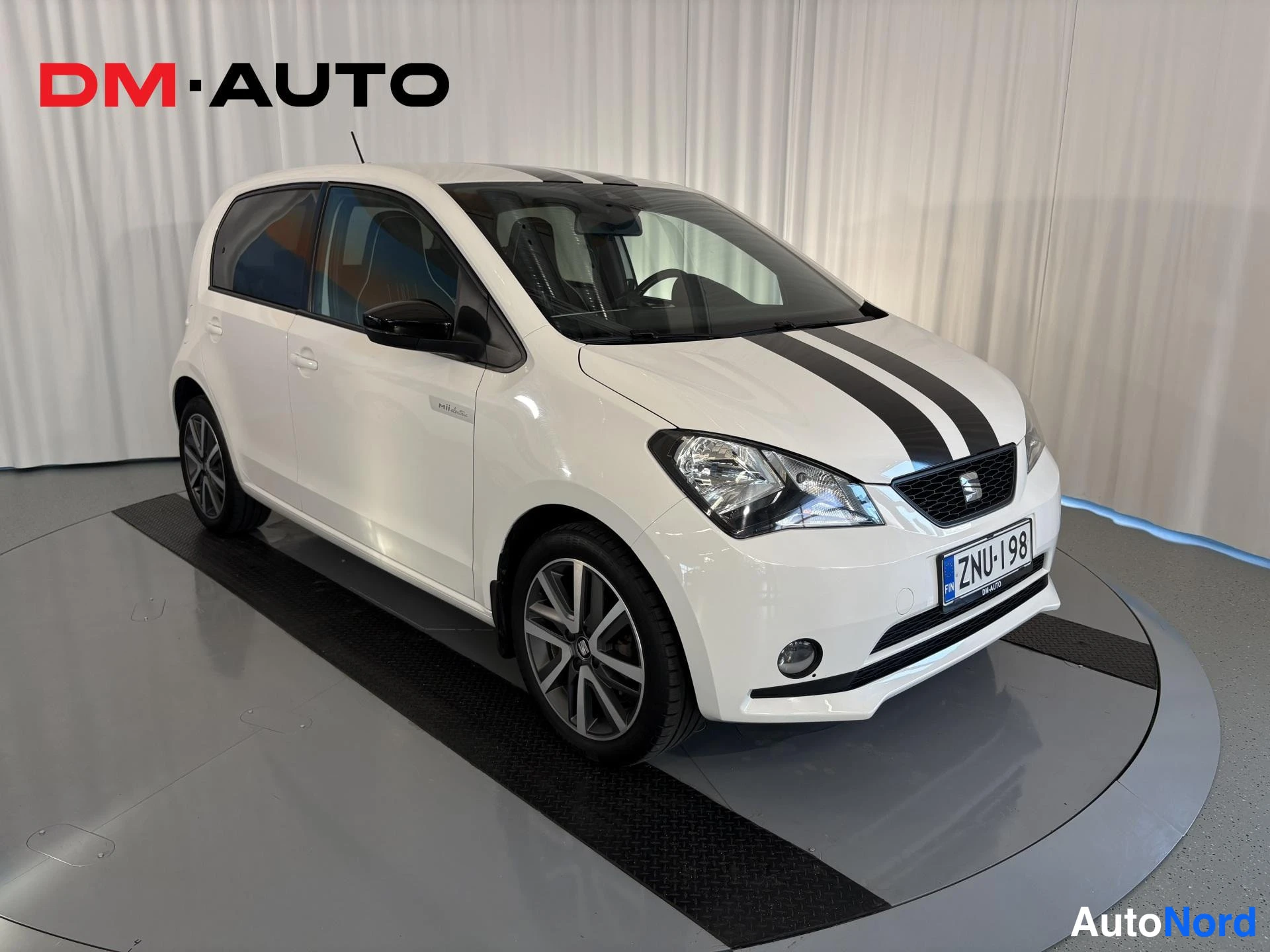 Etuviisto, nokka oikealle: White SEAT Mii Electric photographed from the front-right three-quarter angle: grille and headlight plus the flank are visible, black double stripe on bo