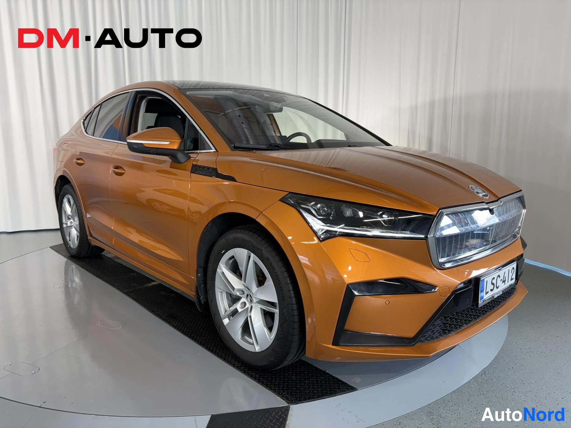Etuviisto, nokka oikealle: Oranssi Skoda Enyaq Coupé kulmaetanäkyvissä keulaa oikealle kääntyneenä, hopeavanteet ja rekisteri.