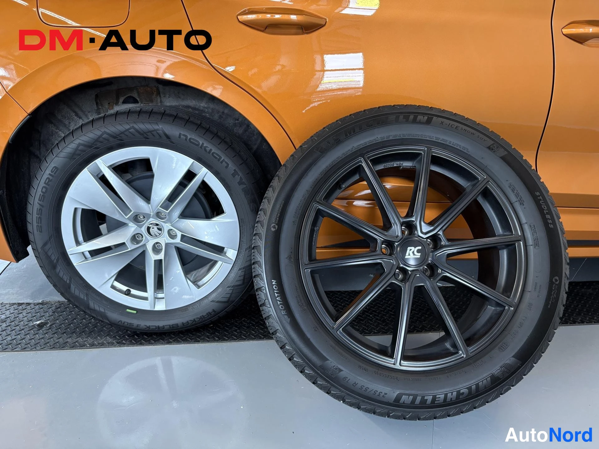 Sivulta: Auton takan pyörä ja vieressä uusi musta MICHELIN-talvirengas vanteella; korin ovipellit oranssit.