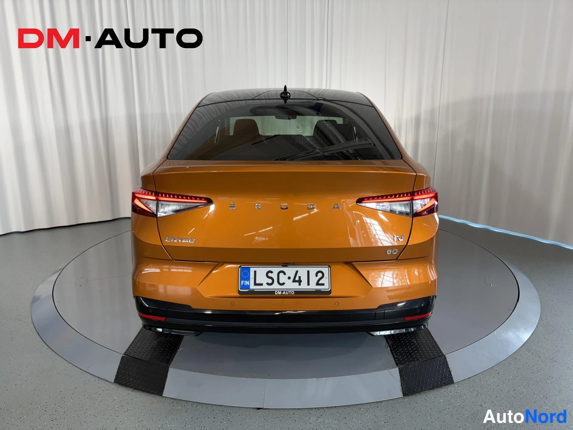 Takaa: Oranssin auton takaosa suoraan takaa, SKODA ENYAQ iV 80 -merkinnät, punaiset takavalot ja rekisterikilpi.