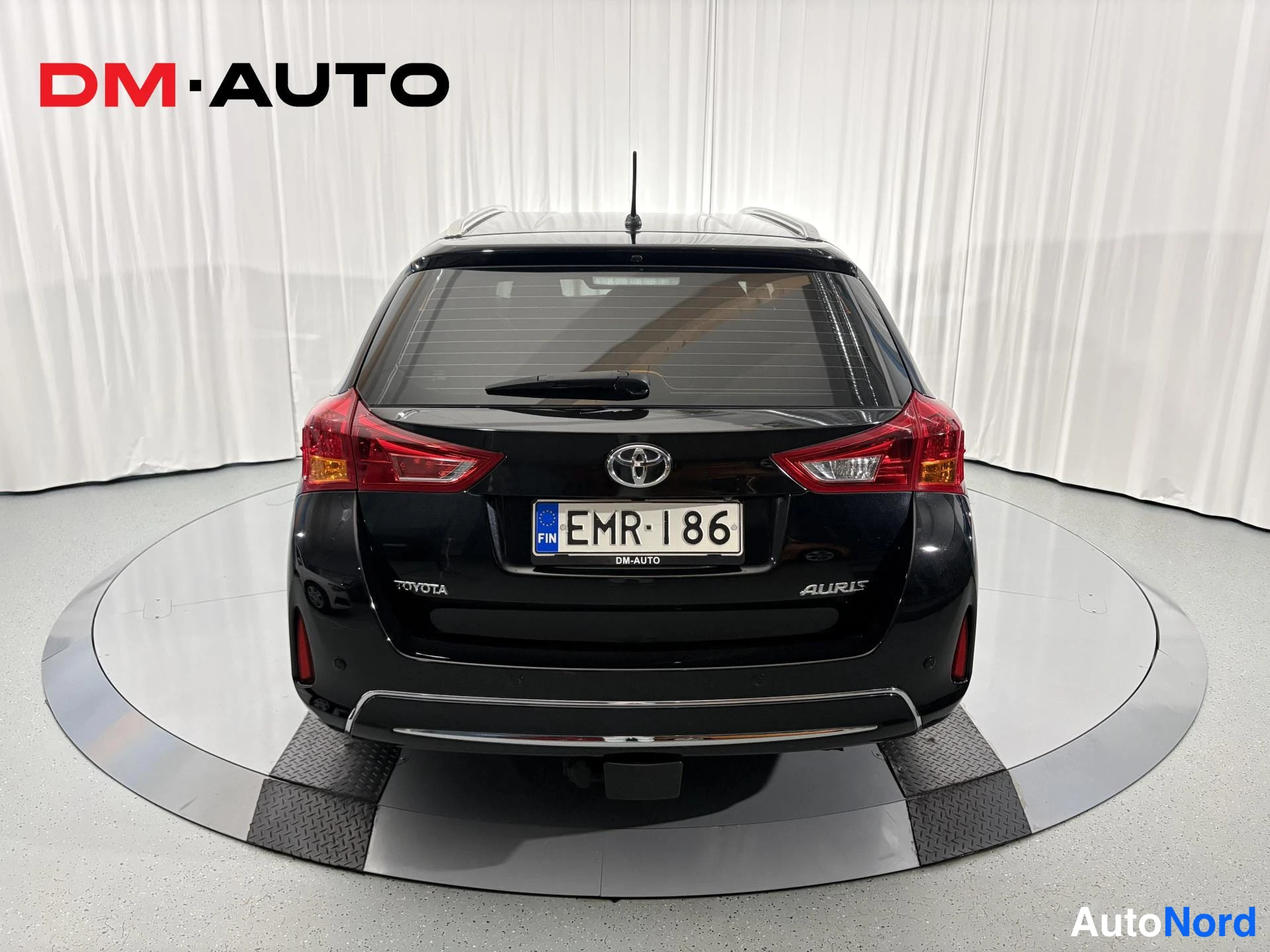 Takaa: Musta farmariauto suoraan takaa kuvattuna, Toyota-merkki ja Auris-kyltti takaluukussa, rekisterikilpi, vetokoulukkeessa takapuskurissa.